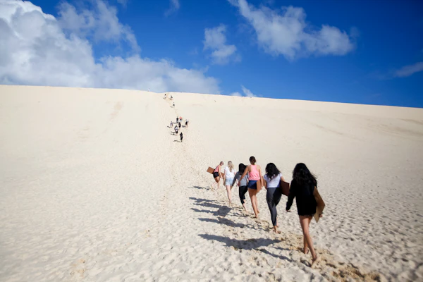 Teen travelers climb in single file up a steep slope of white sand under a blue sky