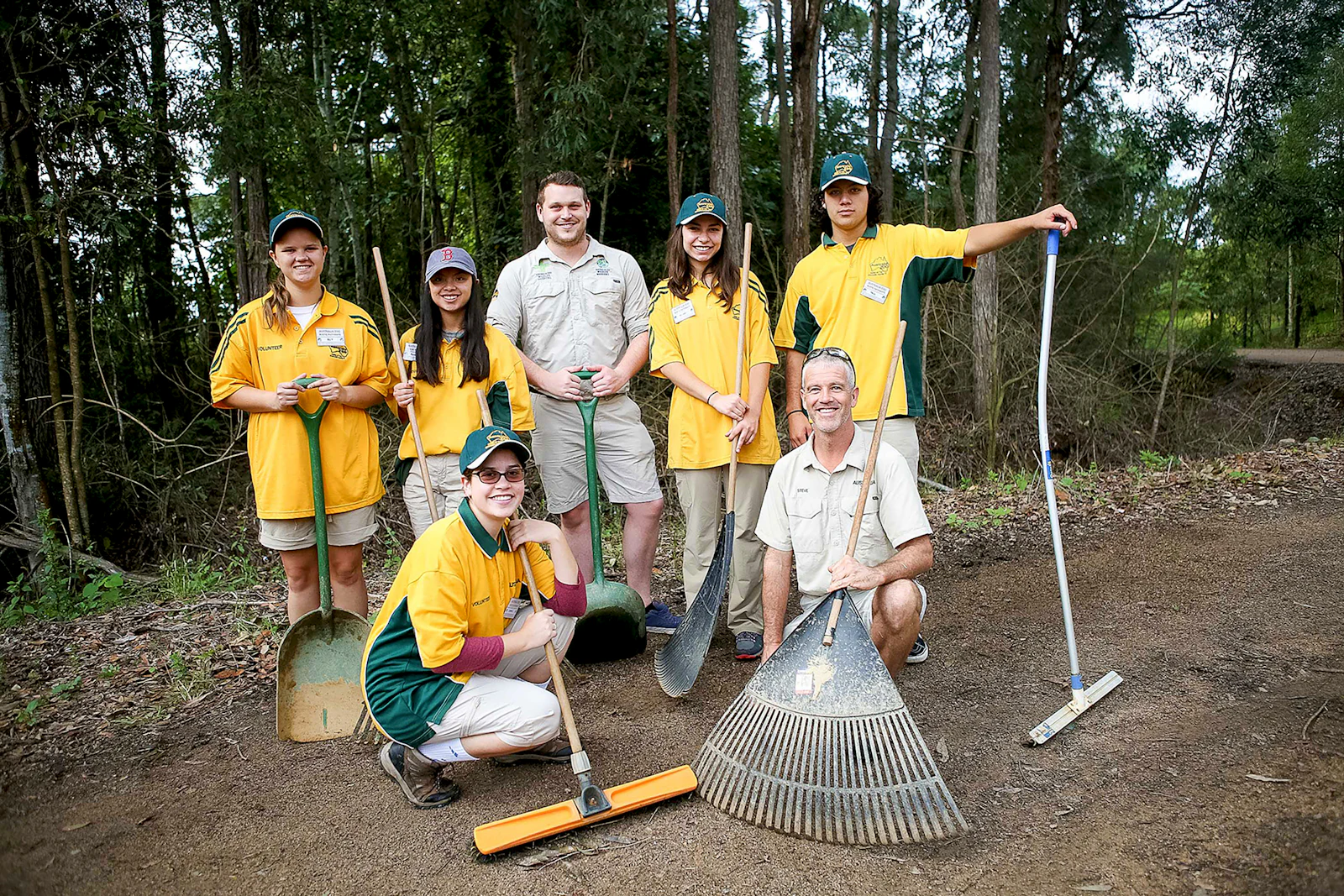 Students in yellow uniform shirts pose with rakes, brooms and shovels beside two adults in a forest clearing
