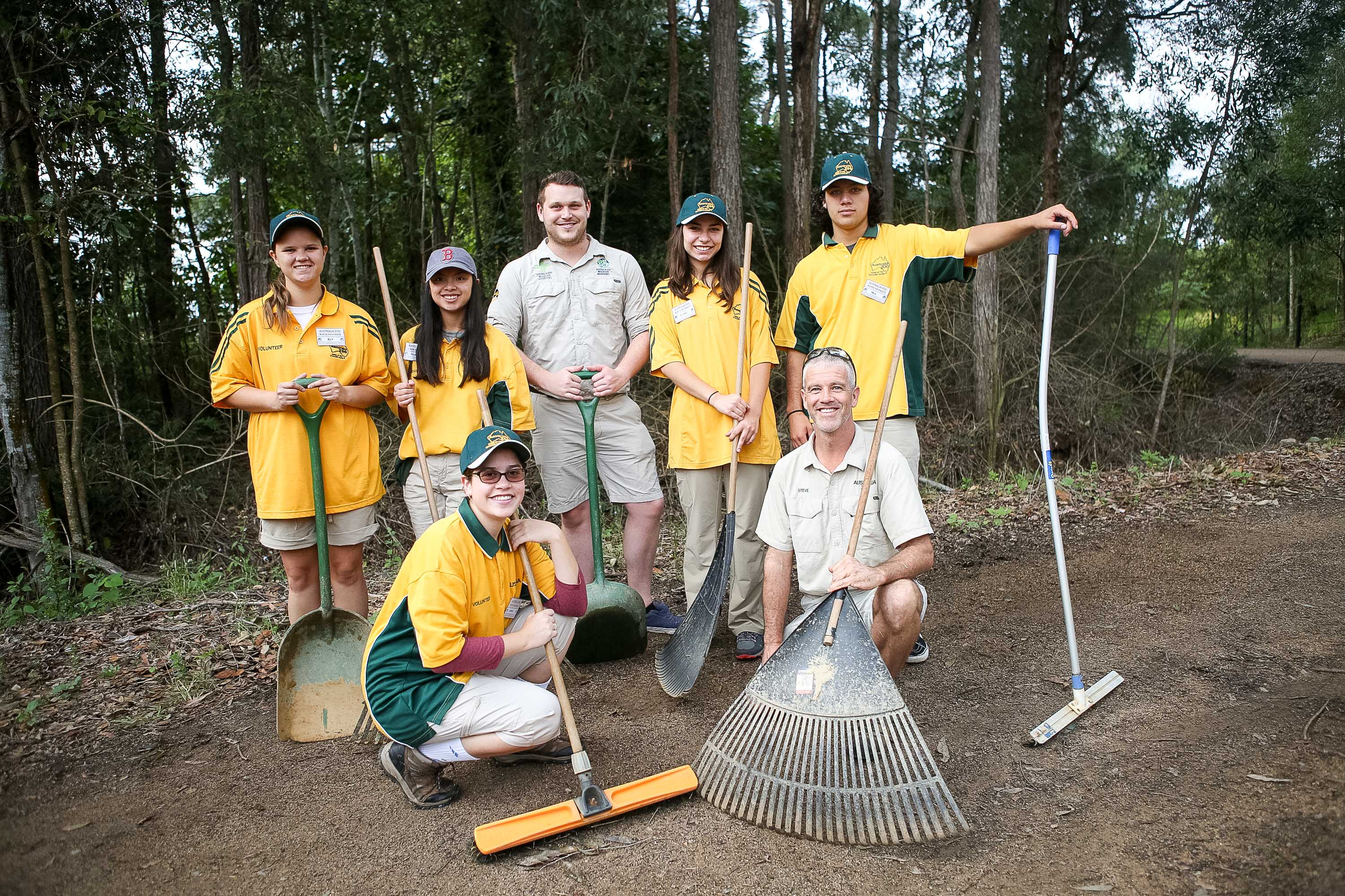 A school trip pauses on a dirt path in the woods, students in matching uniforms posing with rakes and shovels
