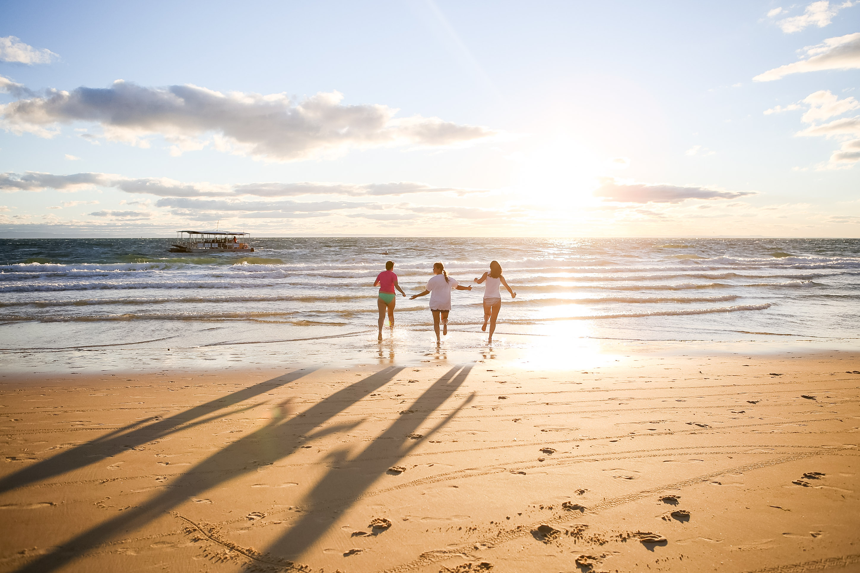 Students hold hands running into the ocean at sunset with a boat offshore