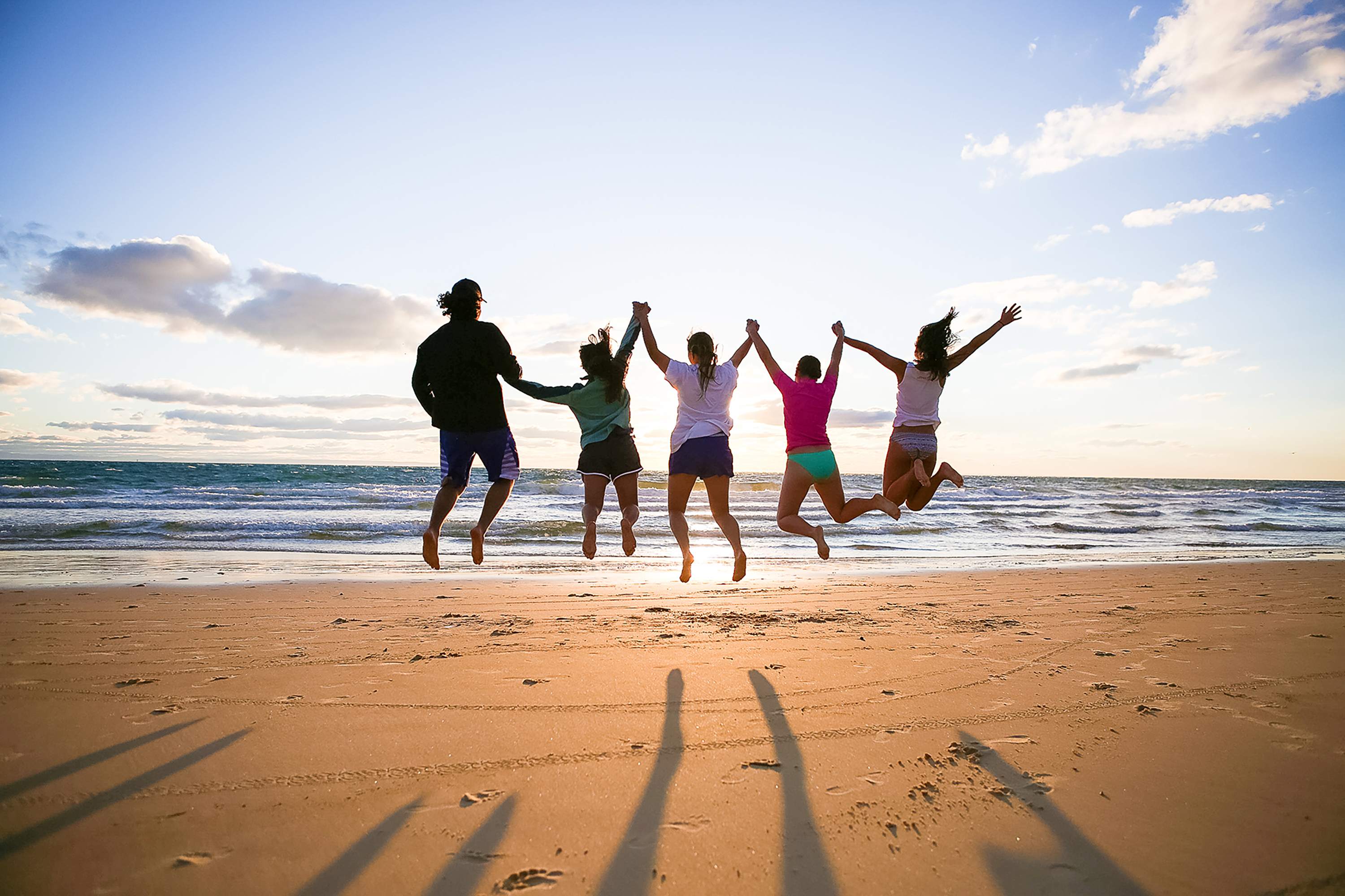 Students hold hands and jump for joy on a beach at sunset