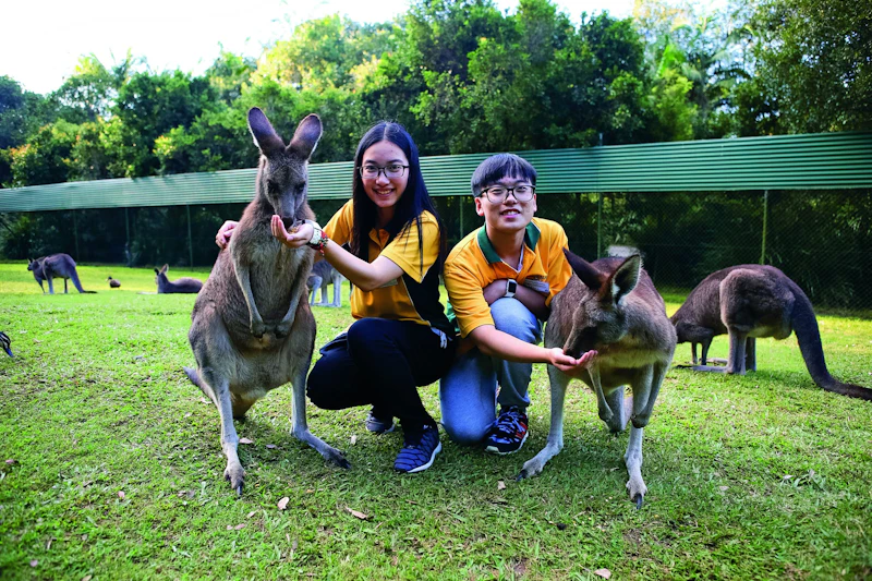 Two teen travelers crouch on the grass between two kangaroos, with more kangaroos grazing behind them in Australia