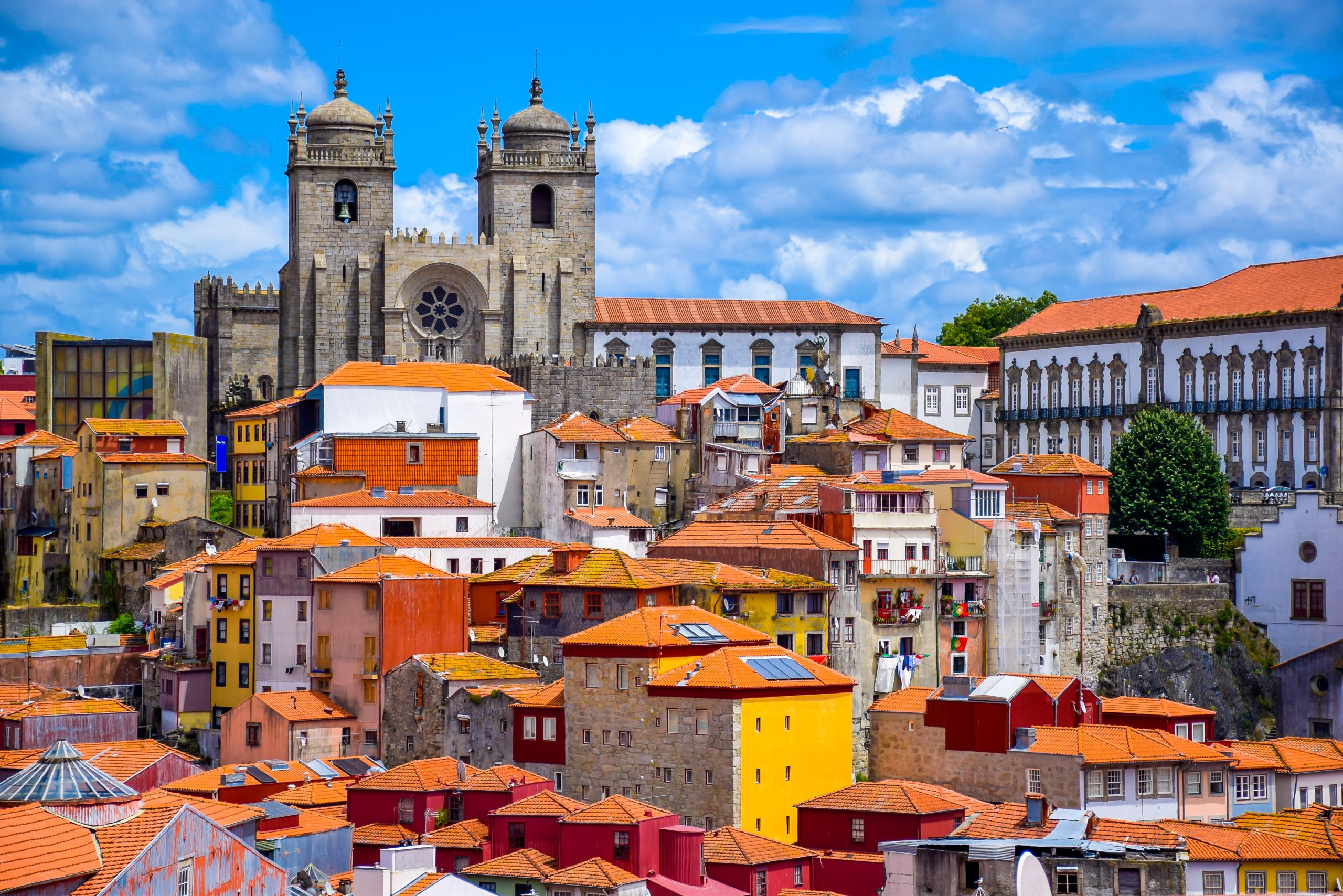 Tightly packed old town houses in ochre, red, blue and white with terracotta roofs climb a slope, and the twin stone bell towers and rose window of a large cathedral rise behind them under a blue sky.