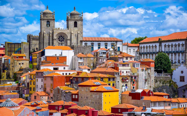 Tightly packed old town houses in ochre, red, blue and white with terracotta roofs climb a slope, and the twin stone bell towers and rose window of a large cathedral rise behind them under a blue sky.