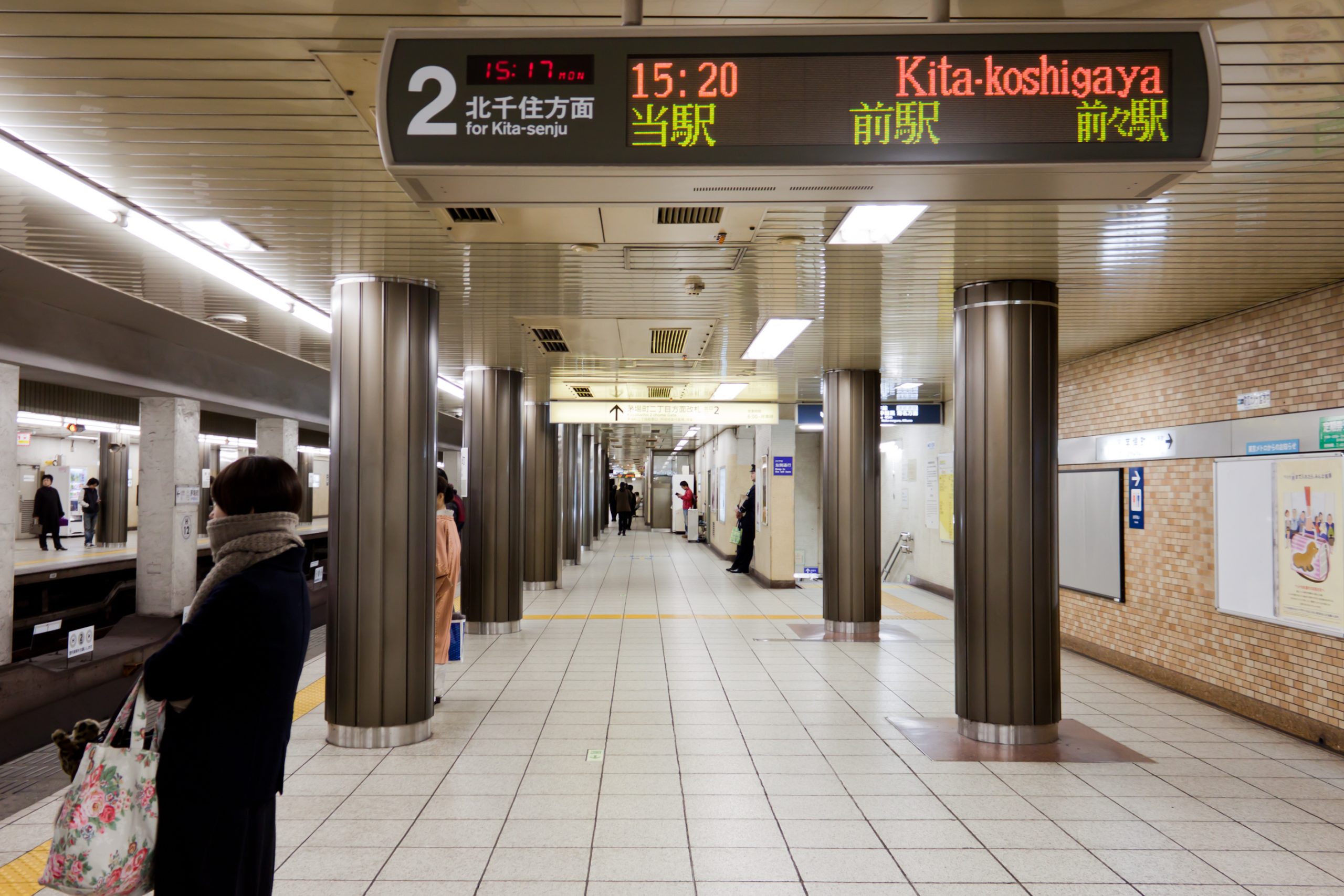 Passengers wait on a Tokyo subway platform beneath a digital train schedule sign