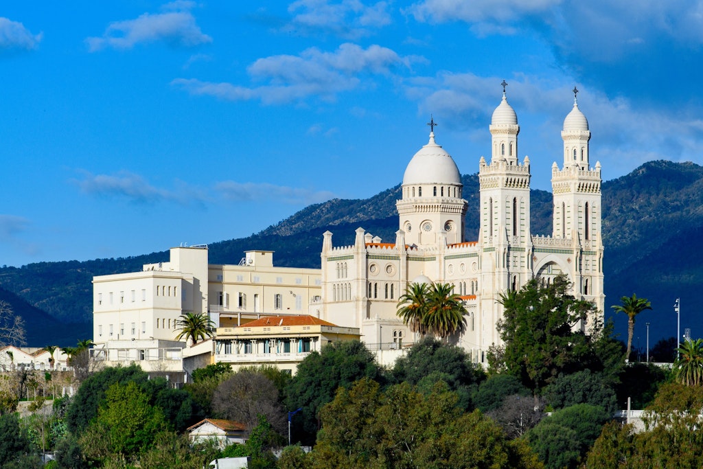 Scenic view of a grand white cathedral with twin towers, set against lush greenery and mountains under a blue sky. Fun facts about algeria