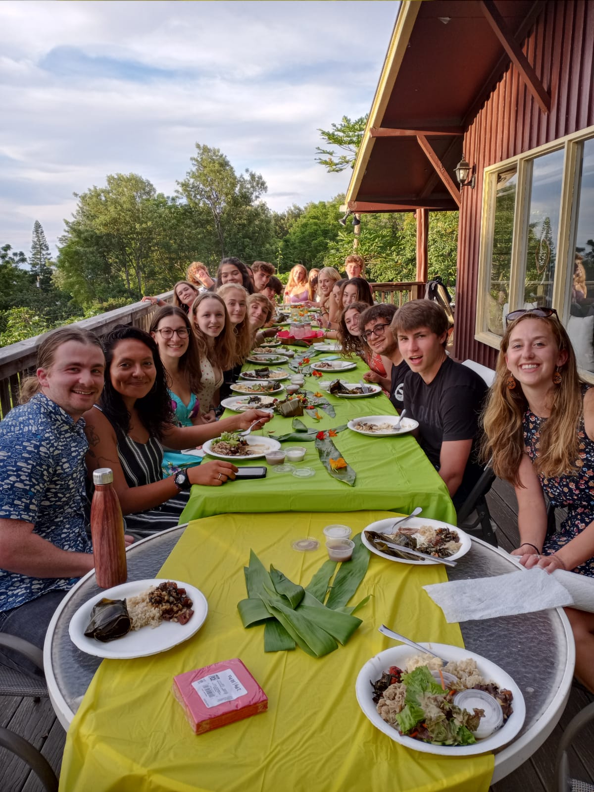 Students share a meal at long tables on an outdoor deck at sunset