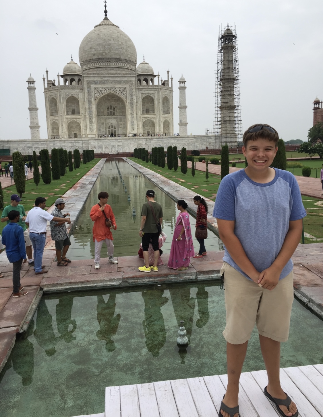 Smiling boy standing in front of the Taj Mahal with tourists in the background.