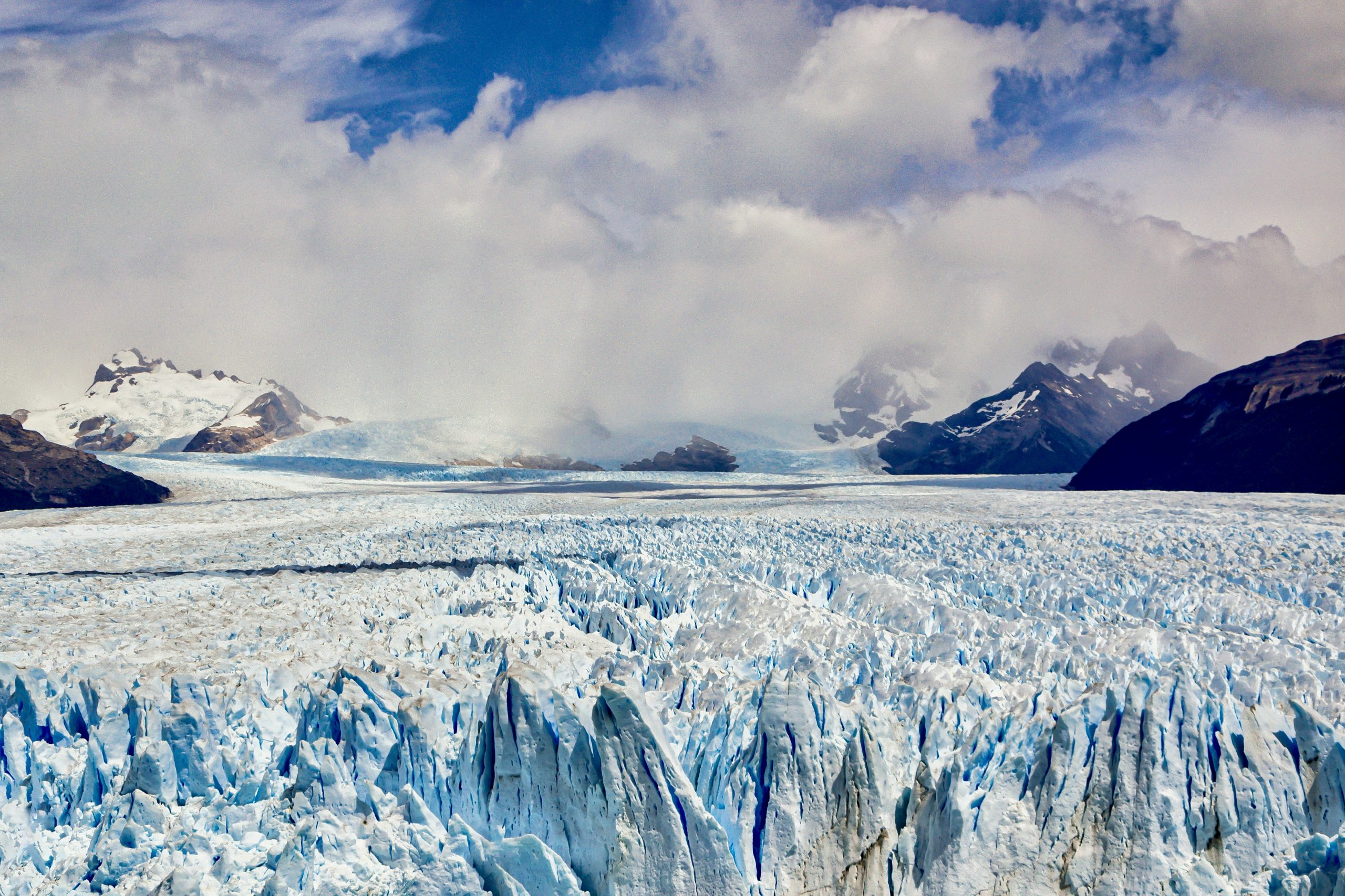 Jagged glacier ice stretches toward snow-capped mountains under a cloudy sky