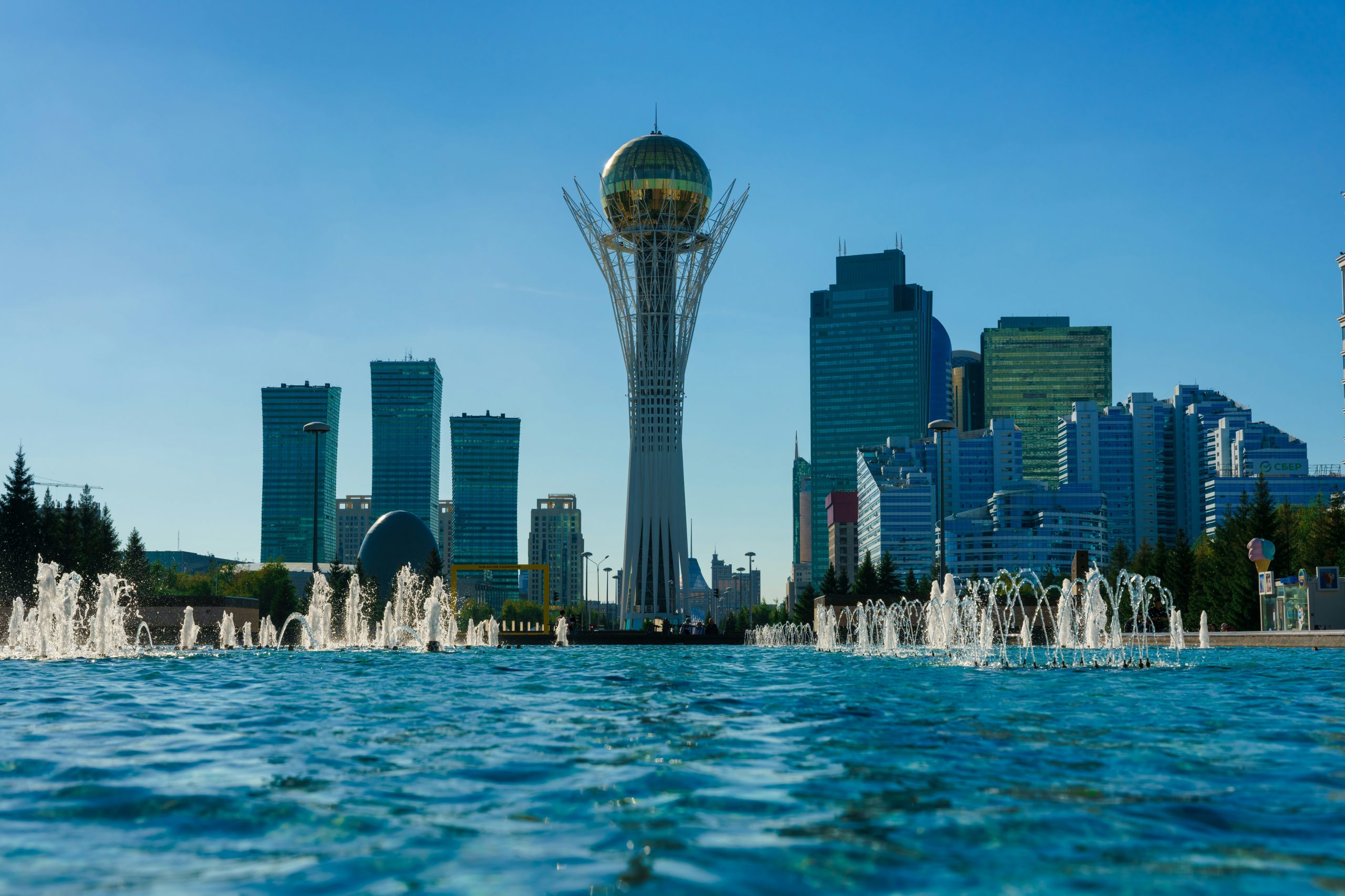 The Baiterek tower's white lattice and gold sphere stand above a fountain pool, with glass office towers on either side under a clear sky.