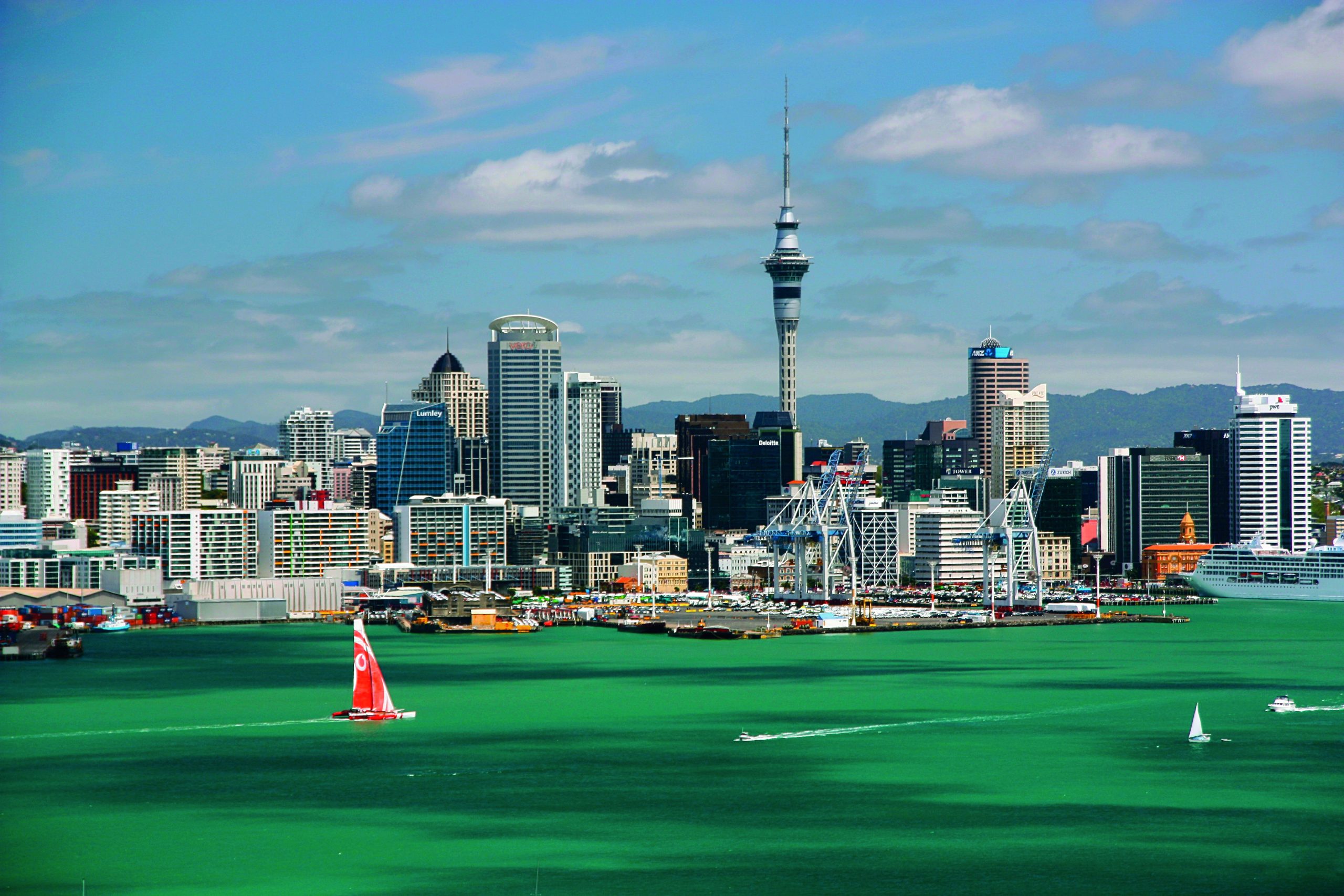 Auckland's harbour with the Sky Tower above the city skyline and a red-sailed yacht on green water.