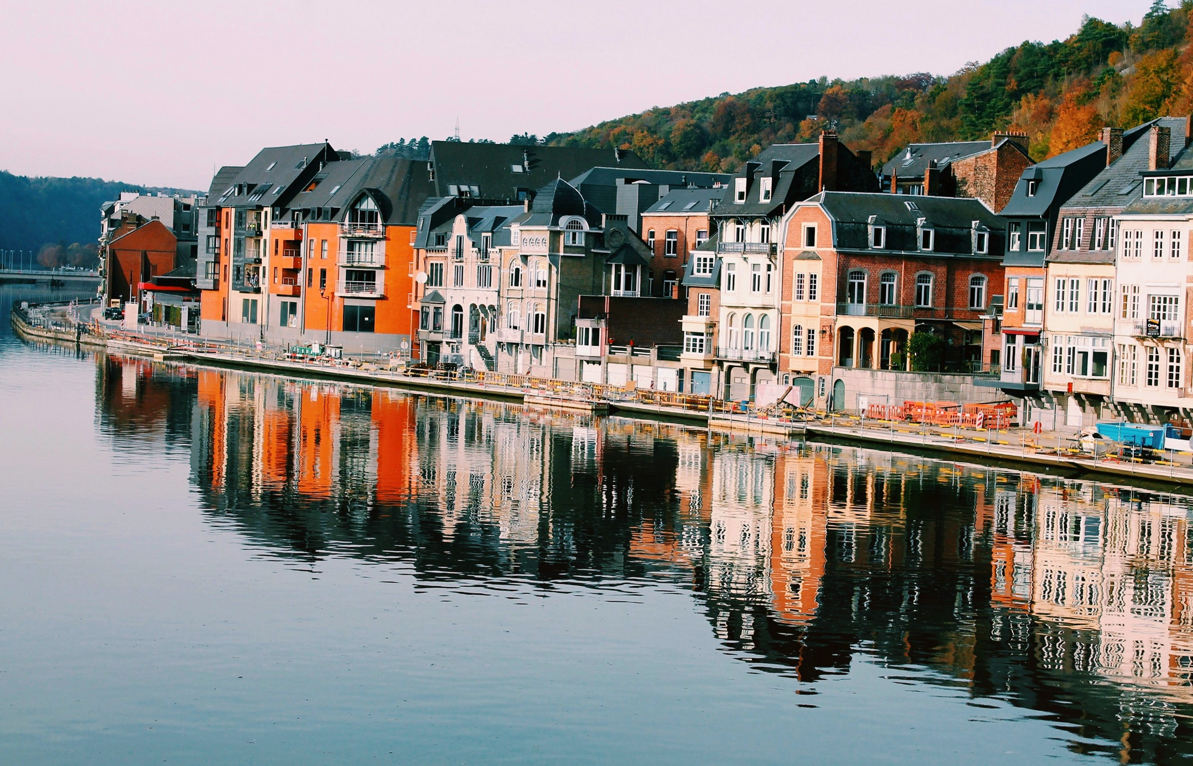 Tall pastel townhouses along a riverside quay, reflected in still water below a wooded ridge.