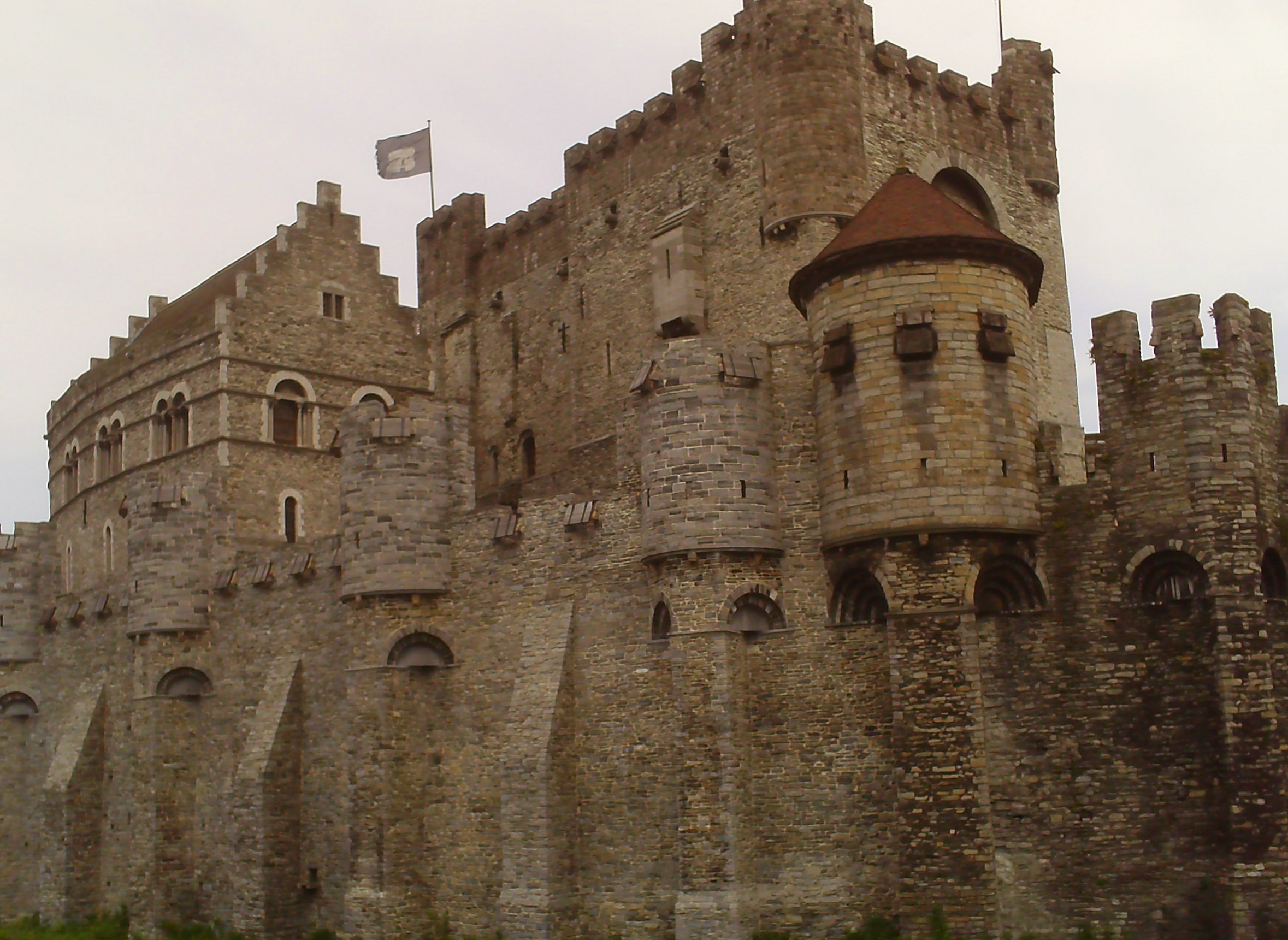 A stone castle with crenellated walls, a round corner turret and a red conical roof, seen from the street.