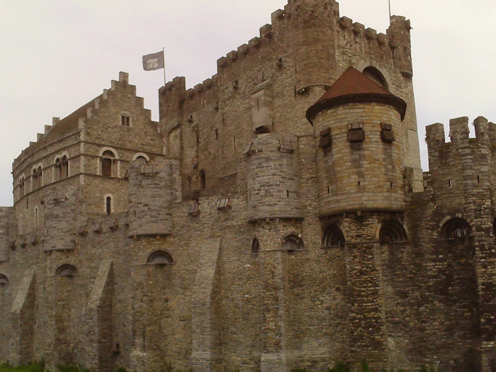A stone castle with crenellated walls, a round corner turret and a red conical roof, seen from the street.