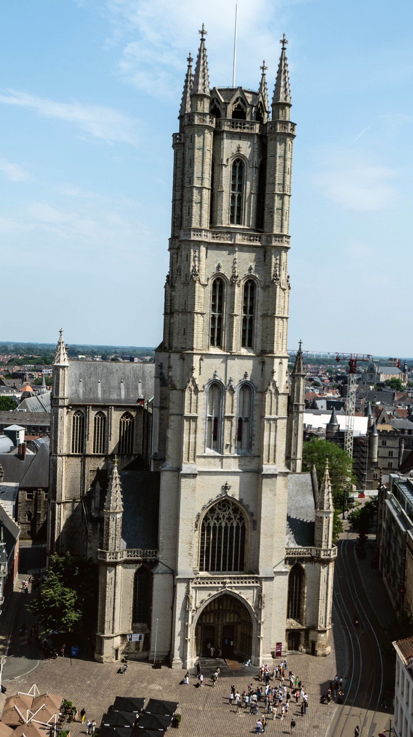 A gothic cathedral tower of pale stone rising above steep rooftops, seen from the height of a neighbouring belfry.