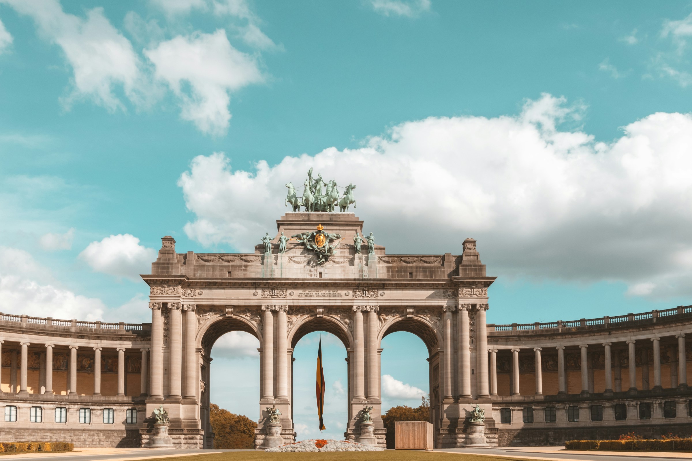 A triumphal arch of three bays topped by a bronze quadriga, flanked by curved colonnades under a bright cloudy sky.