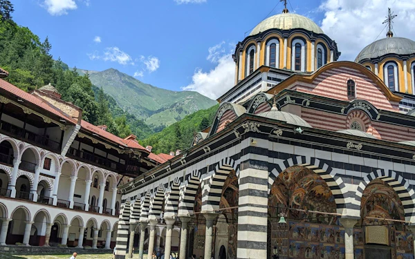 The striped arcades and domed church of Rila Monastery inside its courtyard, with a forested slope behind.