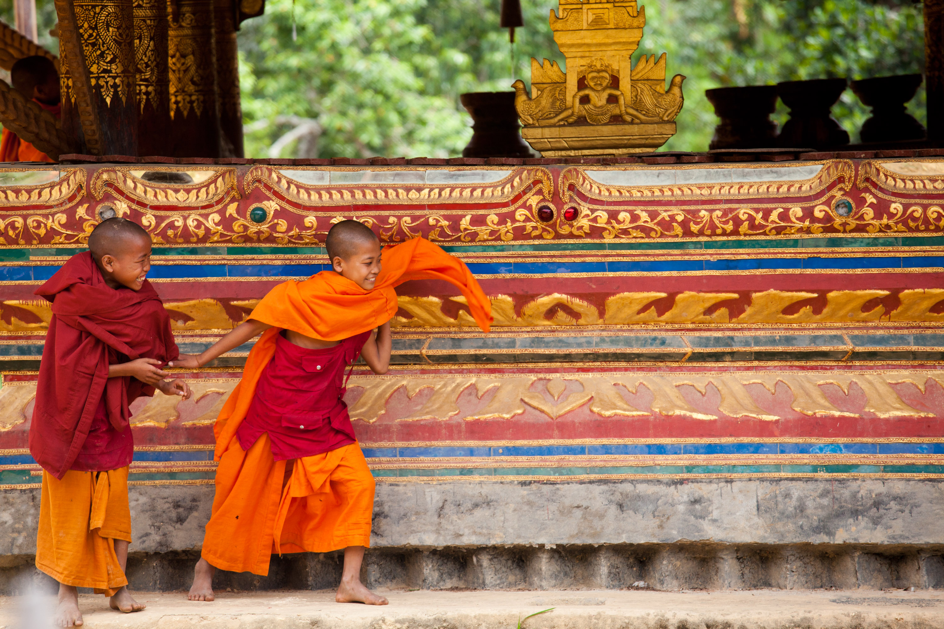 Students watch young monks in robes play alongside an ornately decorated temple wall