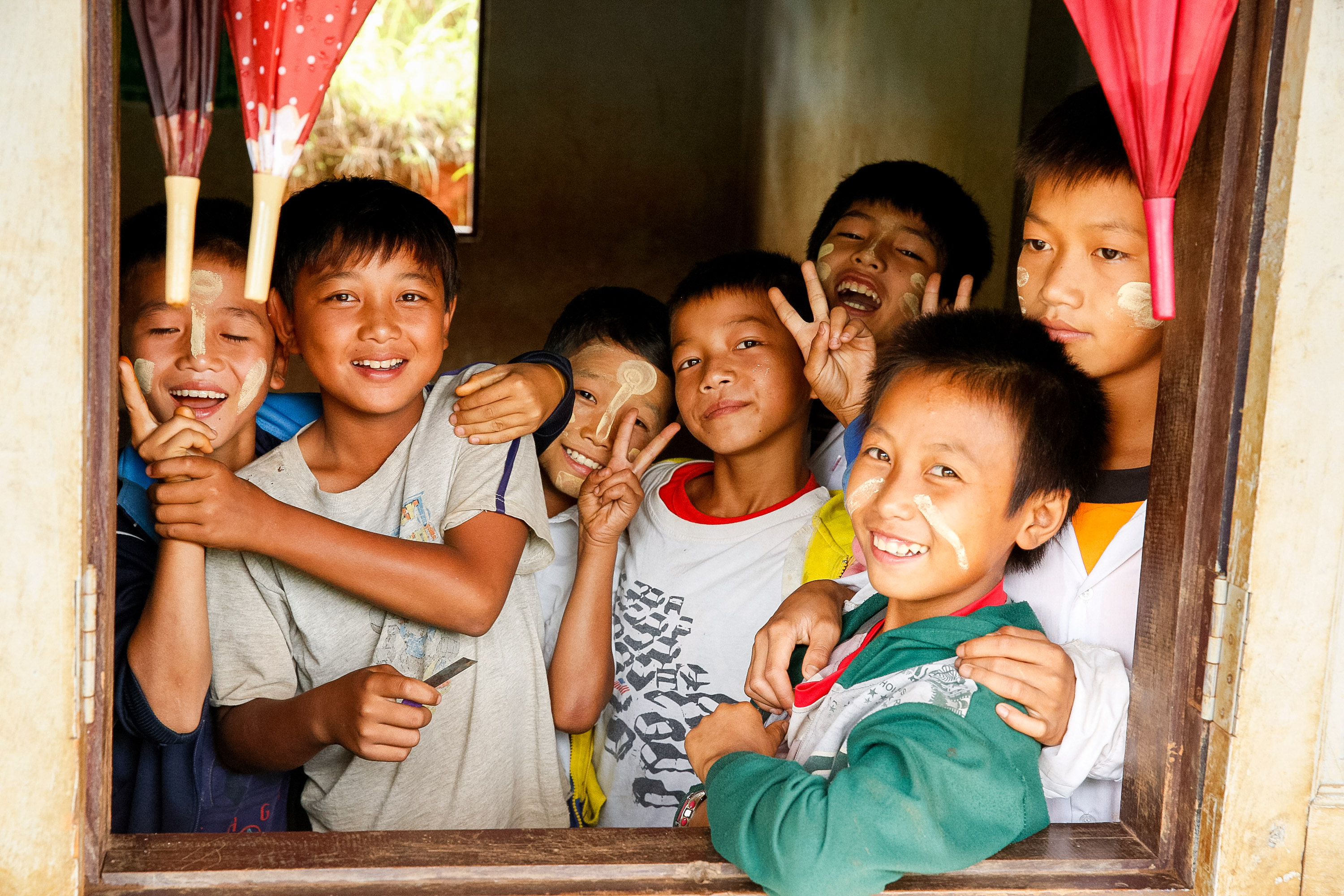 Local children with thanaka paste on their faces smile and pose through a window
