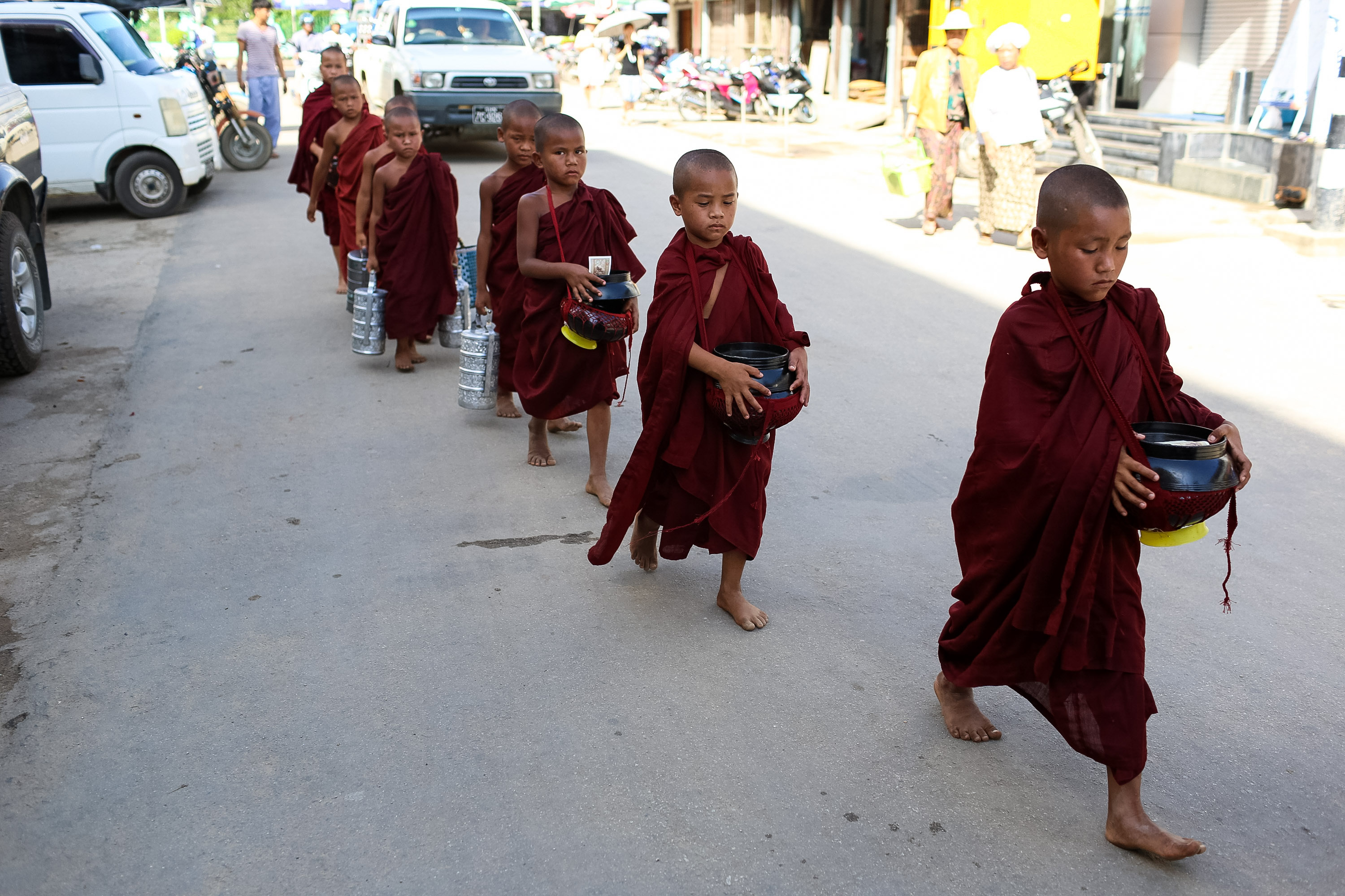 Young Buddhist monks in maroon robes walk barefoot along a street carrying alms bowls
