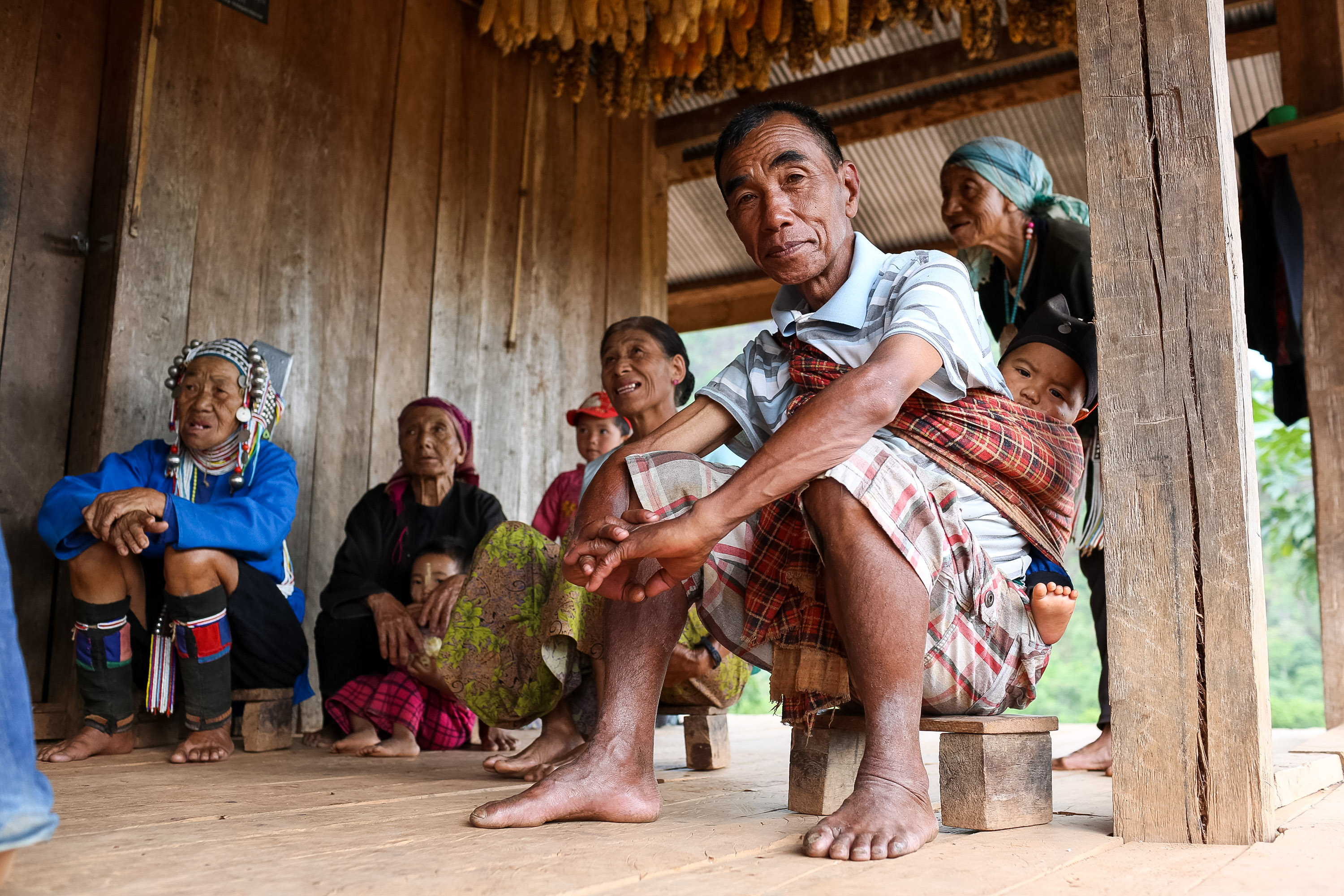 A local family gathers under a wooden stilt house with dried corn hanging above