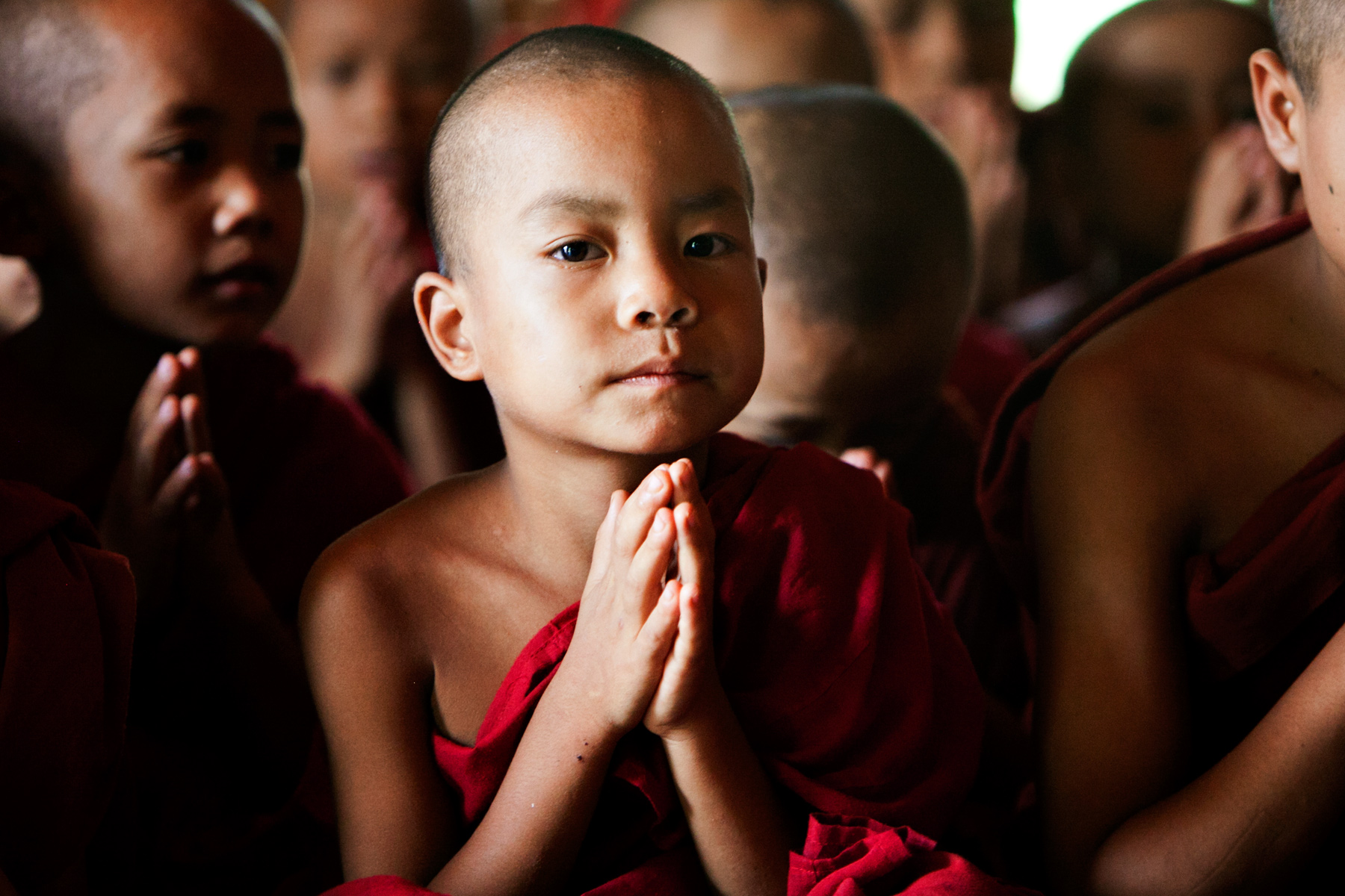Young monk in red robes presses palms together in prayer among fellow novices