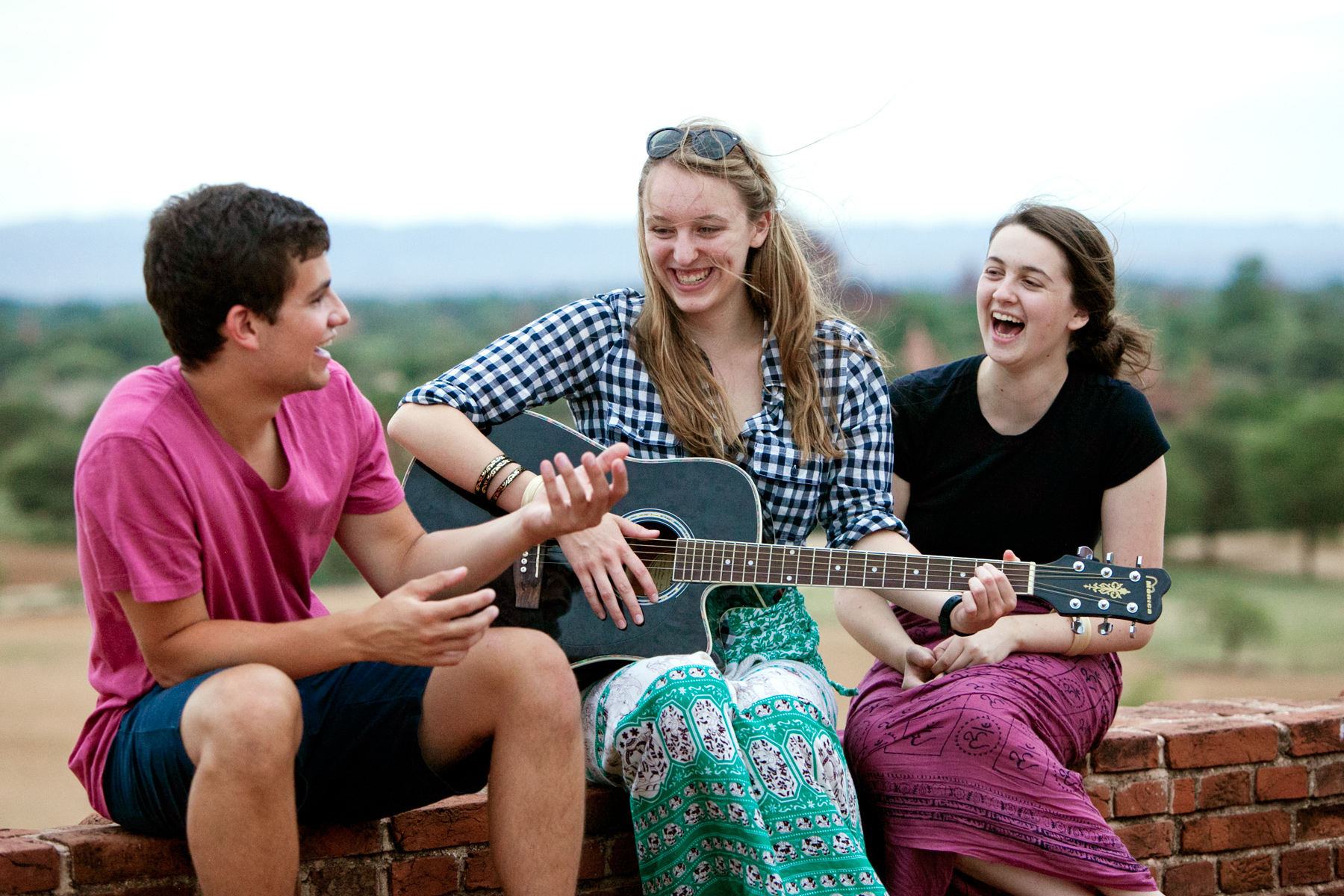 Students laugh together while playing guitar on a brick wall overlooking the countryside