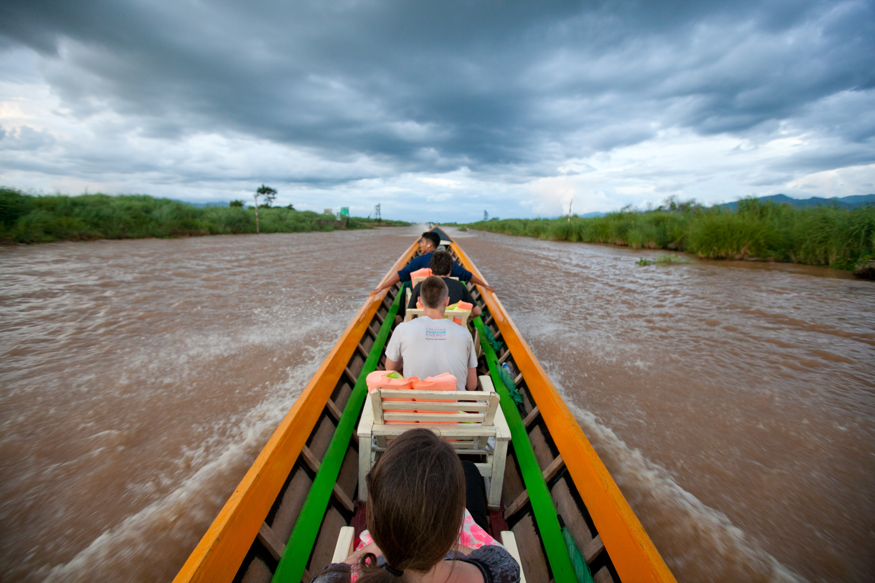 Students ride a long wooden boat down a river under cloudy skies