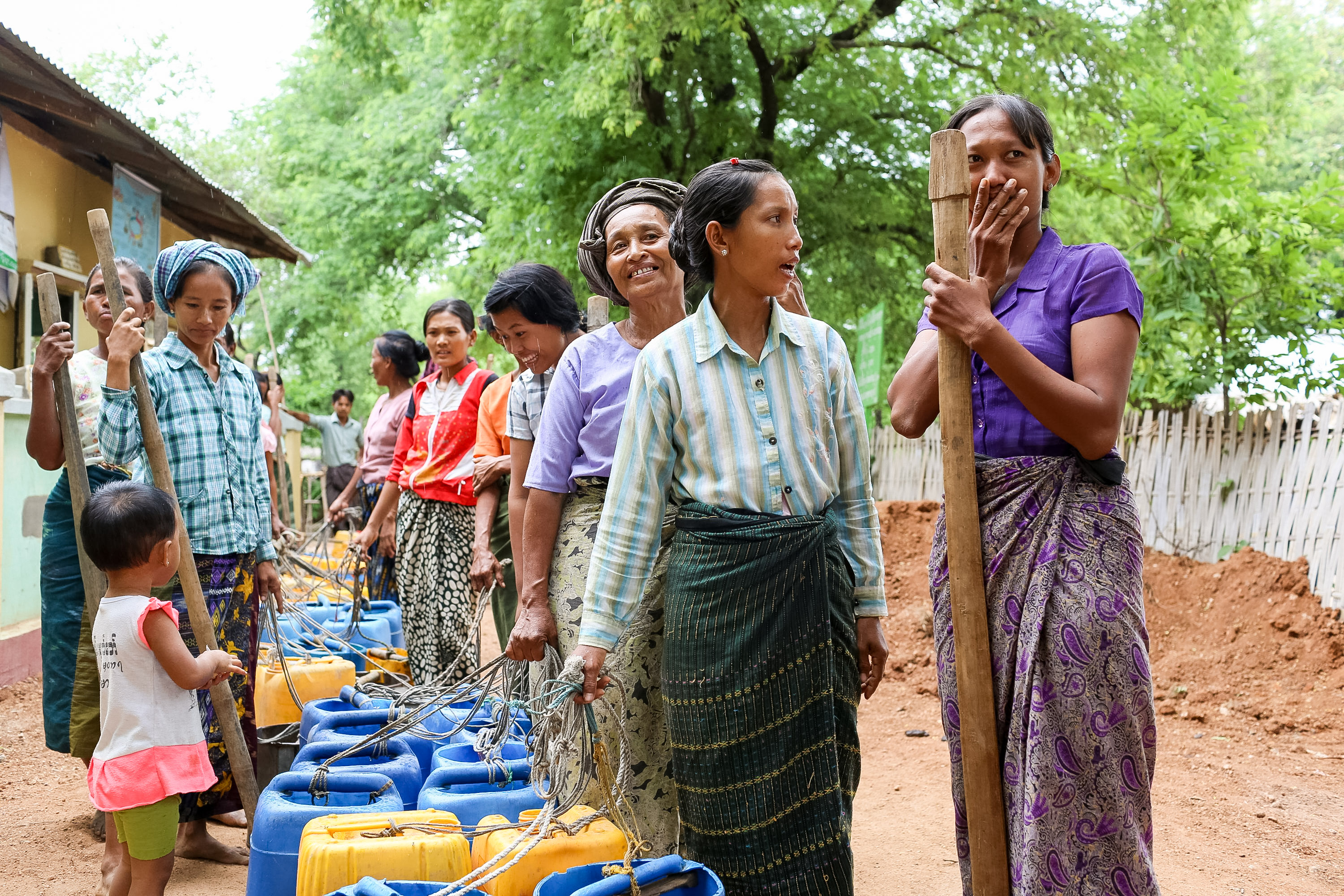 Villagers gather with blue water jugs and long wooden poles on a dirt path