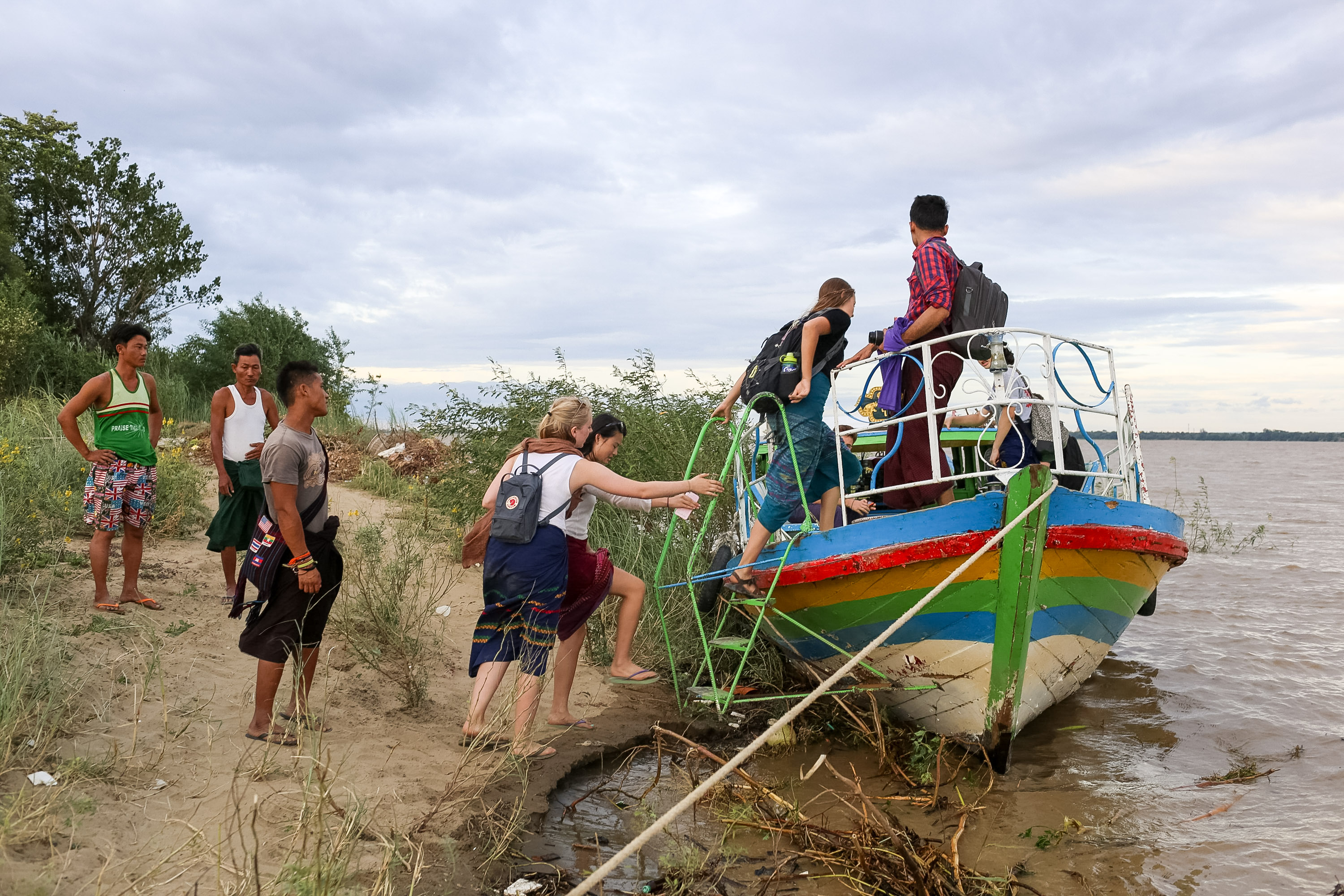 Students climb a ladder into a colorful boat on a riverbank