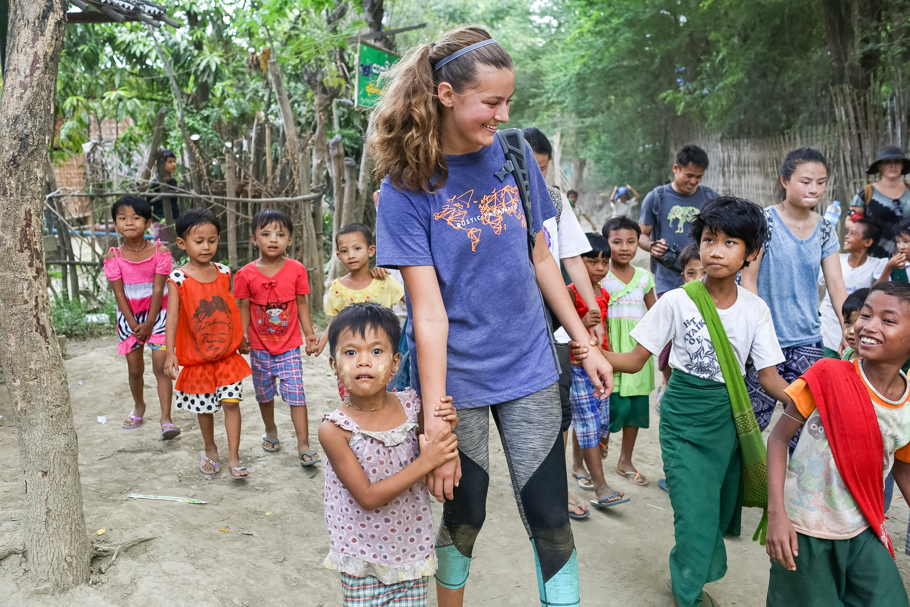 A student walks hand in hand with village children along a dirt path