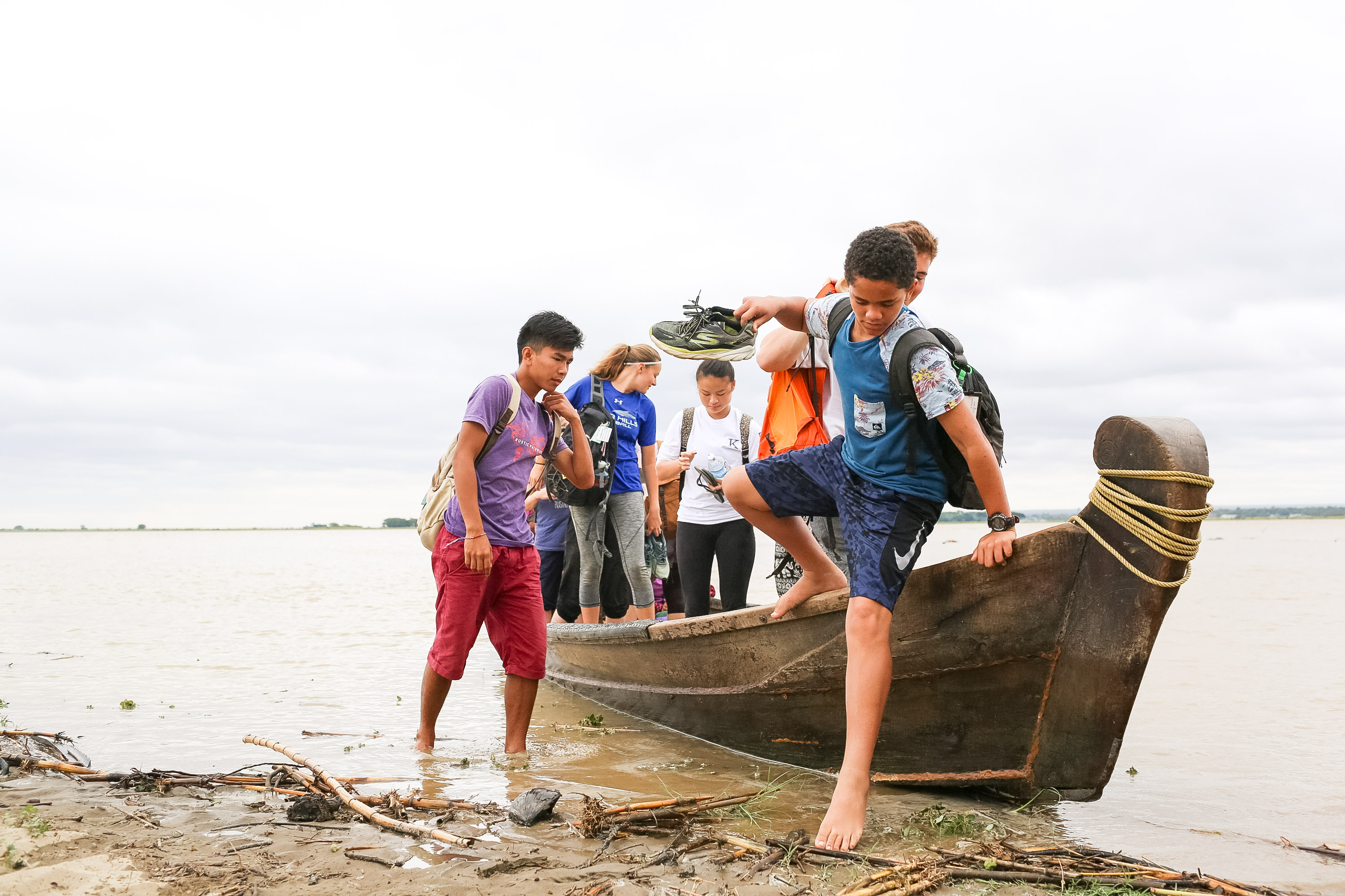Students step off a wooden boat onto a muddy riverbank in Myanmar
