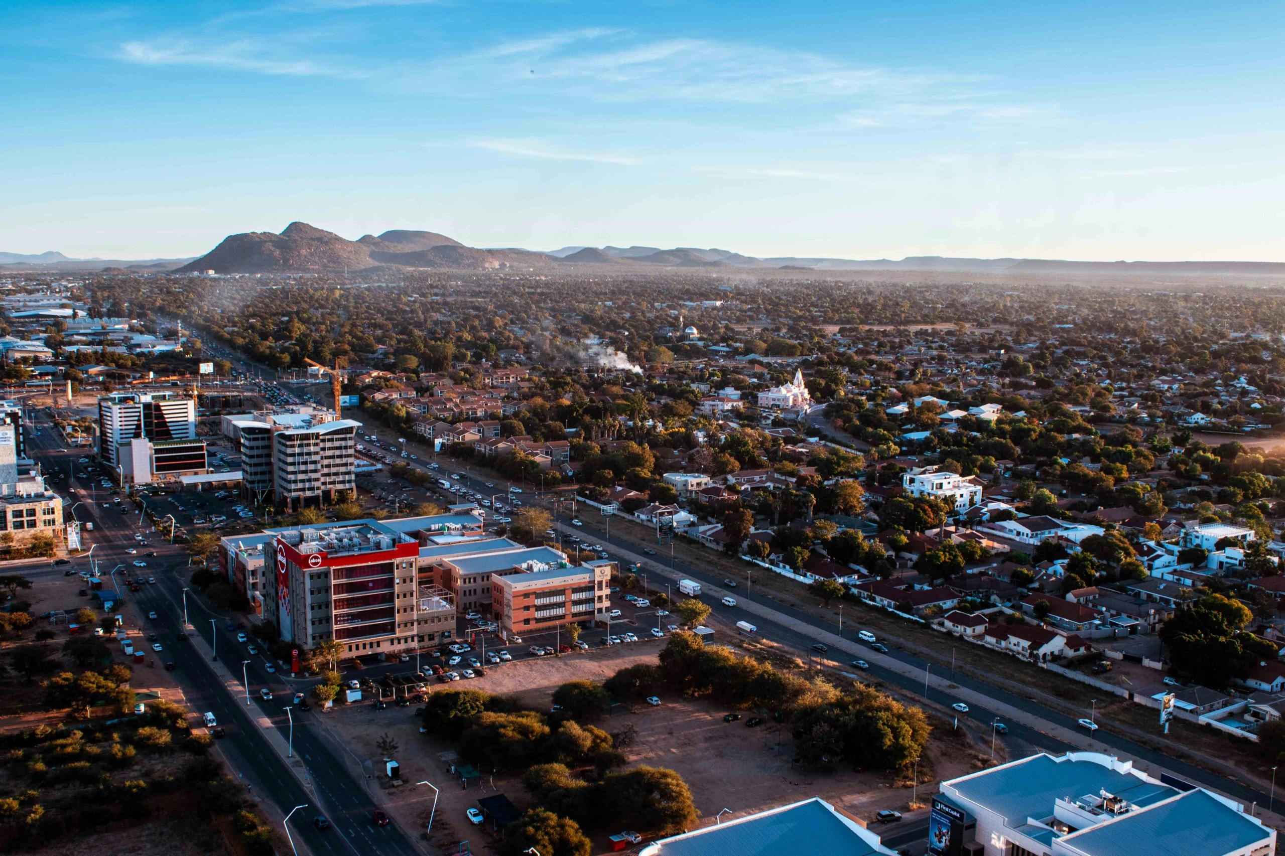 An aerial view of a city's low-rise center and tree-lined suburbs at dusk, with a line of hills on the horizon.