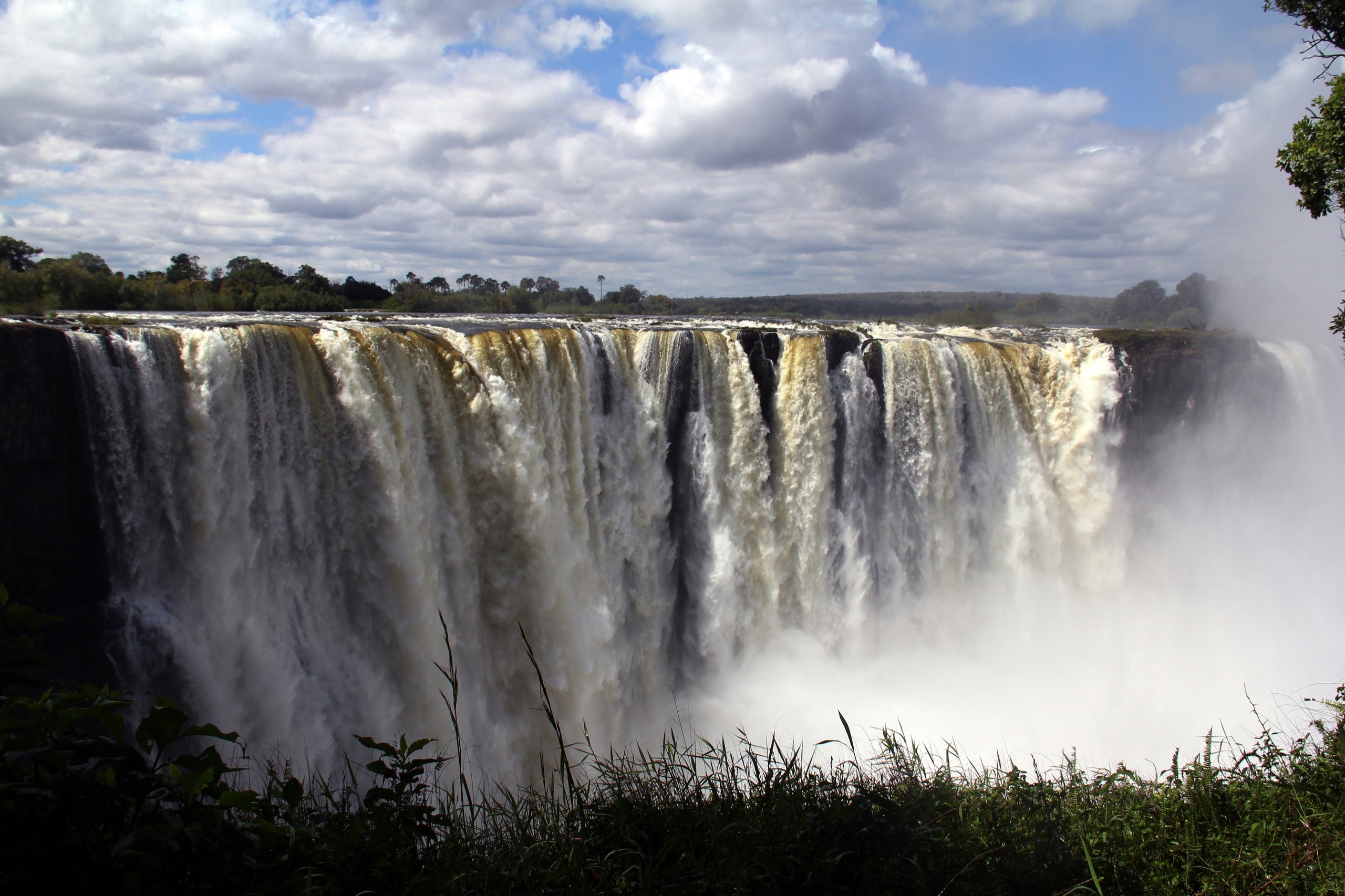 A wide waterfall drops in several channels over a dark cliff into a deep gorge, spray rising against green forest on both sides.