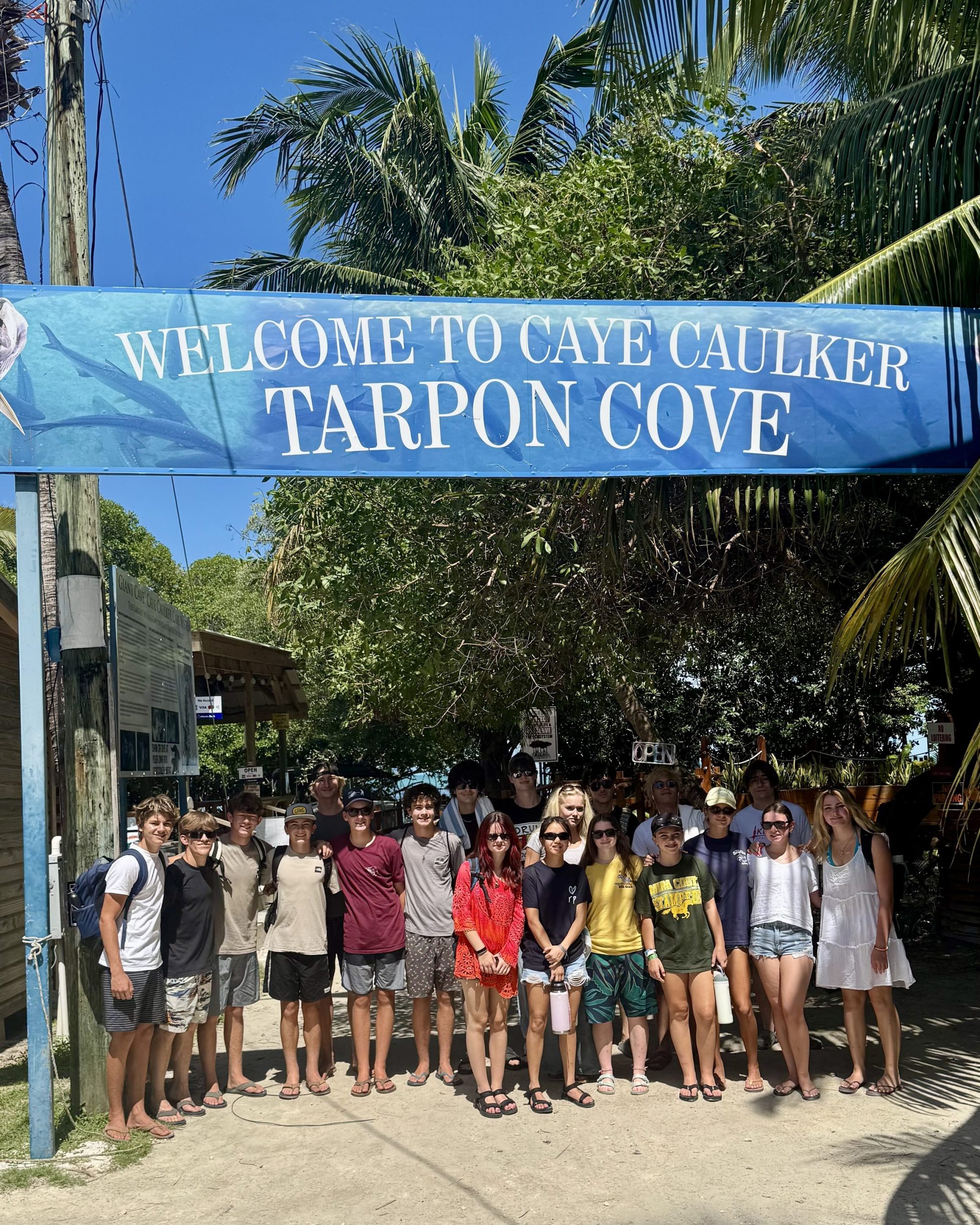 A school group stands together under the Welcome to Caye Caulker Tarpon Cove arch in Belize.