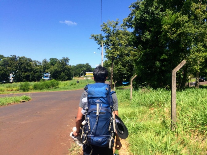 A backpacker facing away from the camera with a blue backpacked on. He is standing in a remote rode somewhere in a remote area of Brazil.