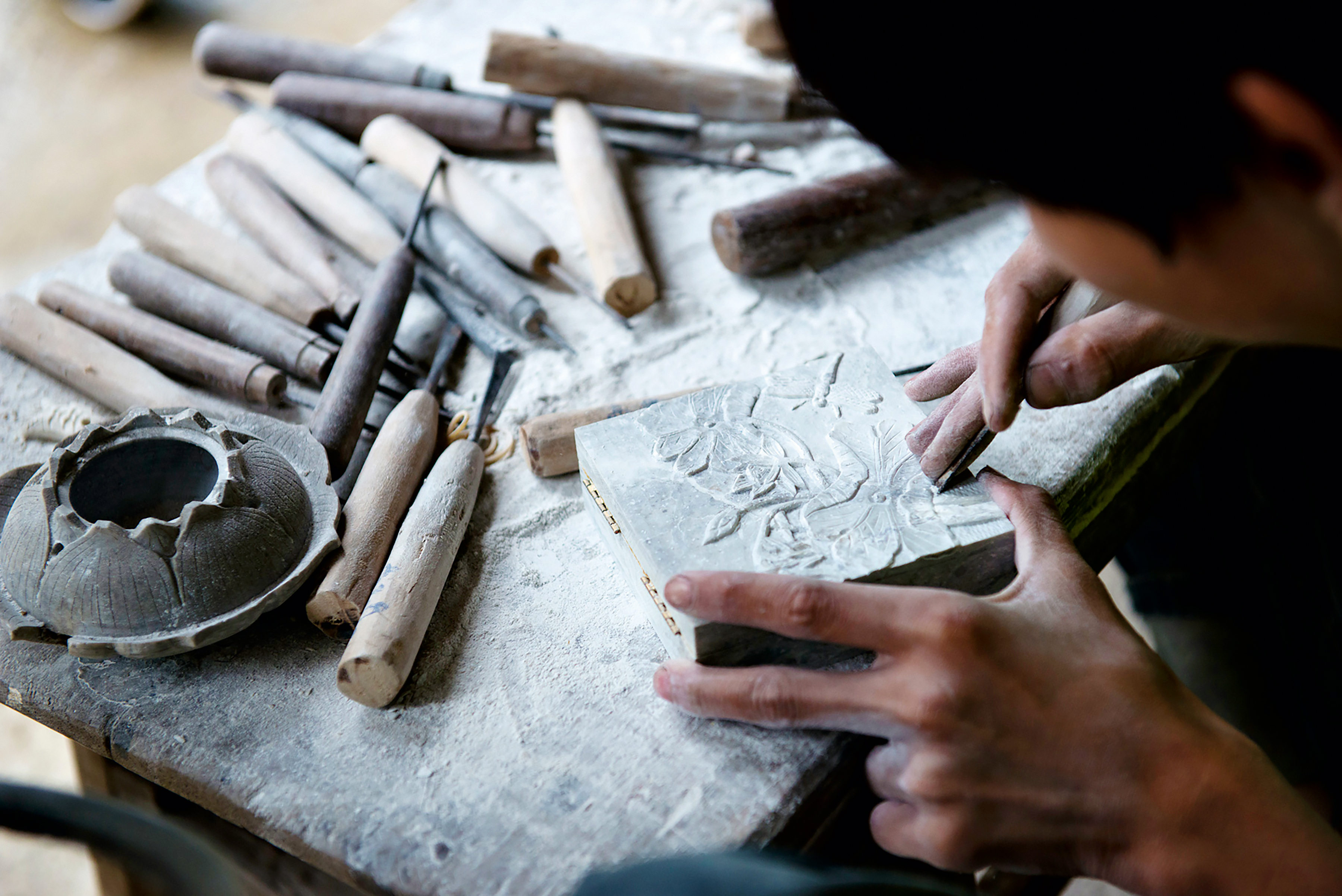 A student carves detailed patterns into a wooden block surrounded by carving tools