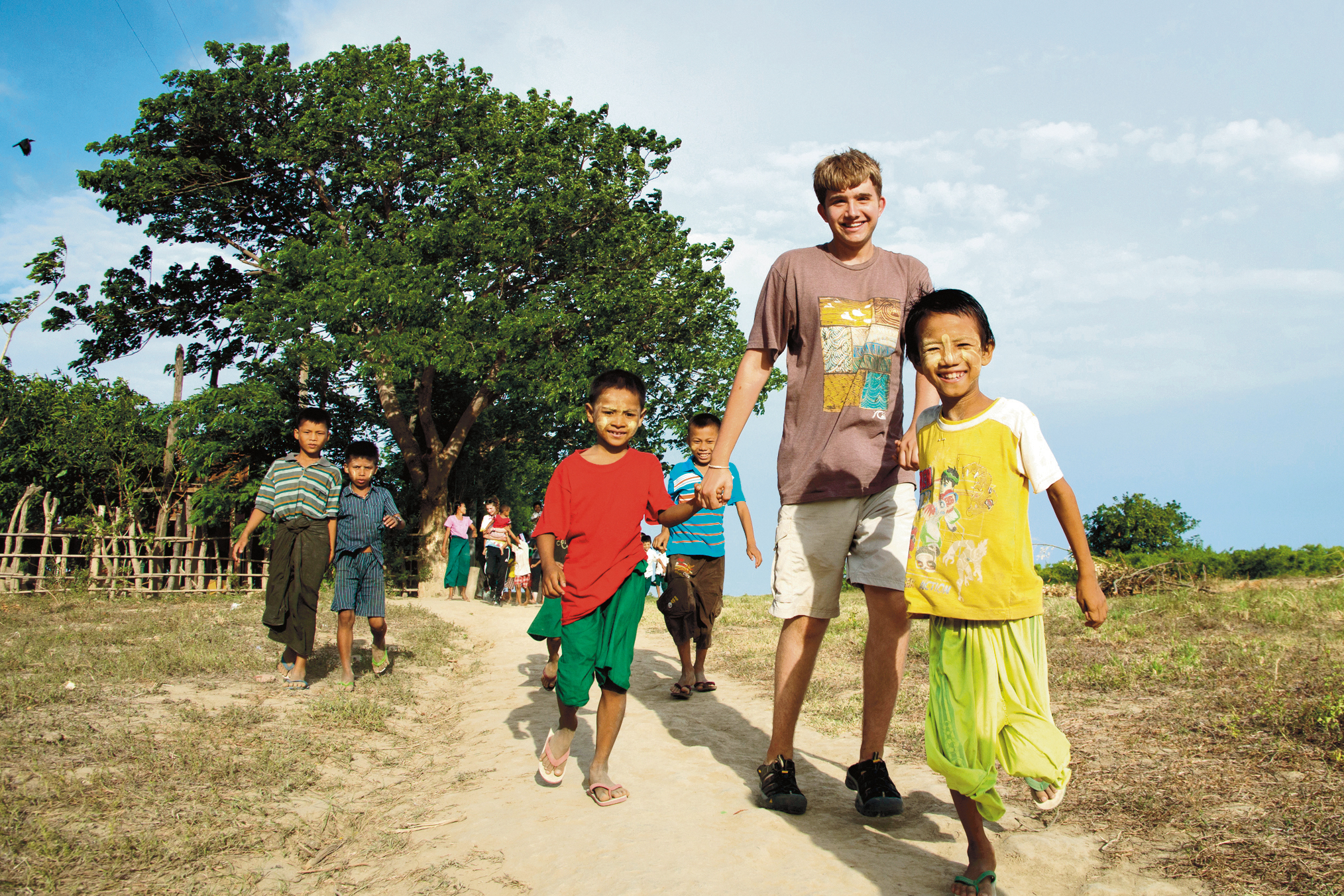 Students walk a dirt path with local children under a large tree