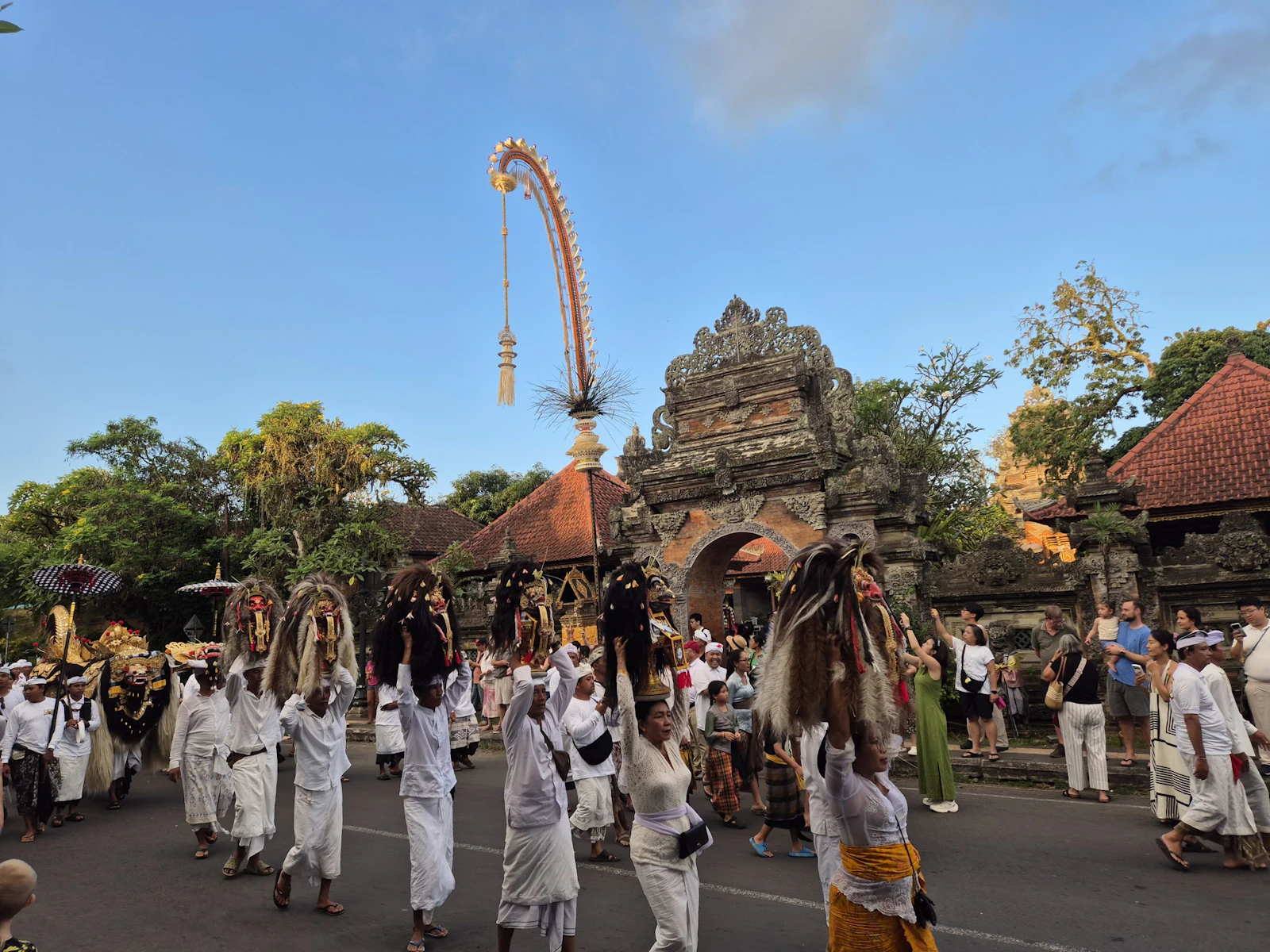 A procession of people in white ceremonial dress passes a carved stone temple gate in Ubud, Bali