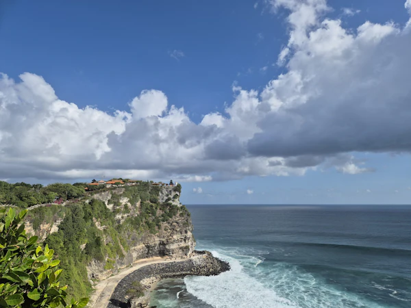 A sheer limestone cliff drops to rough blue sea at Uluwatu, Bali, under a bright sky and big white clouds