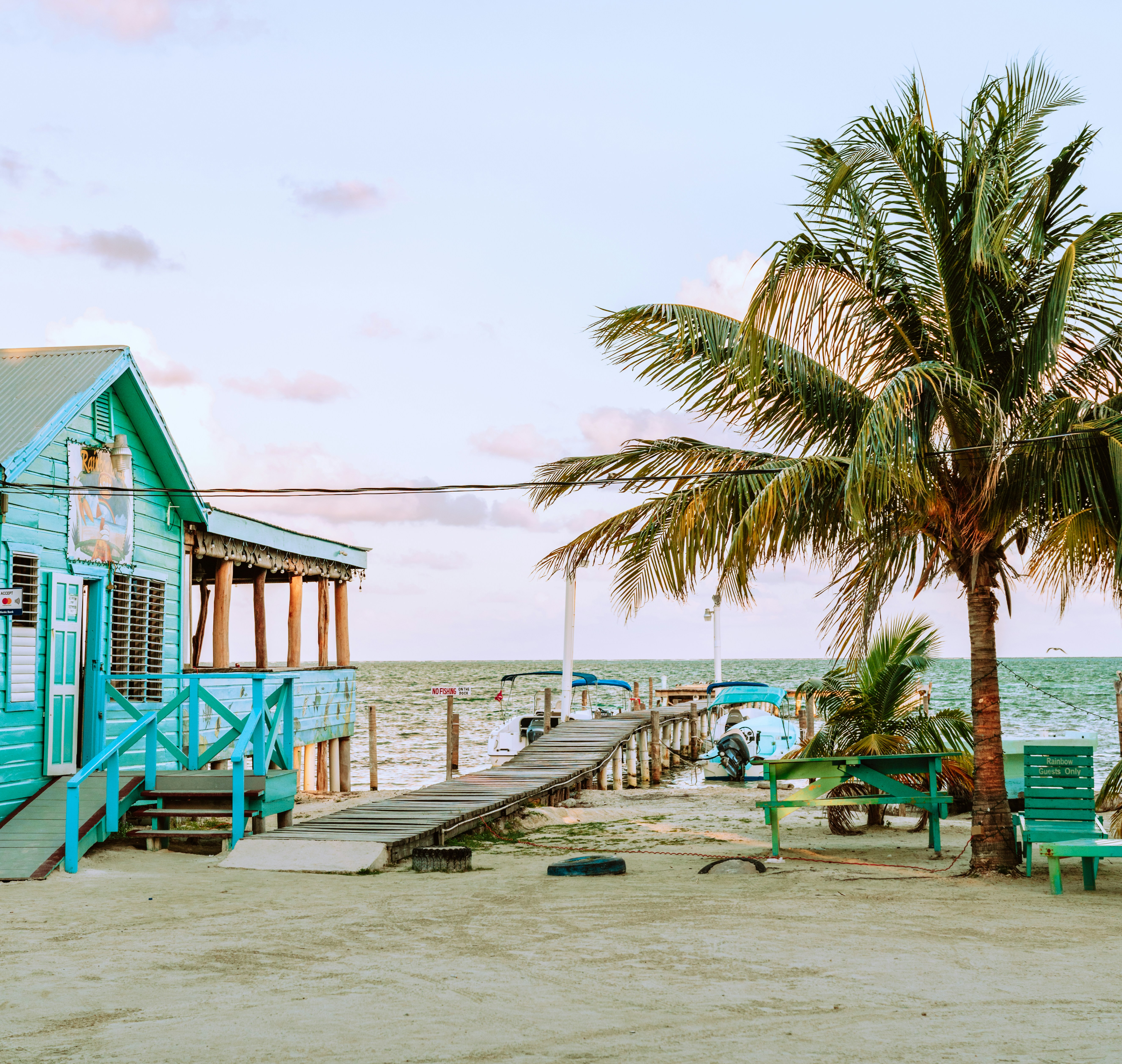 Students walk past a turquoise wooden building with a dock and palm tree along a Belize beach