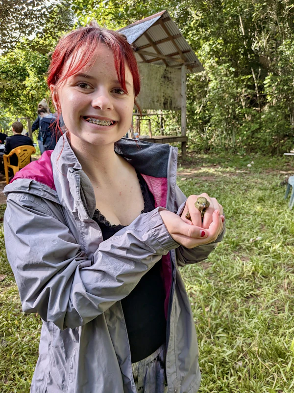 A teen traveler in a grey rain jacket smiles while cupping a small frog in both hands at a jungle field site in Belize