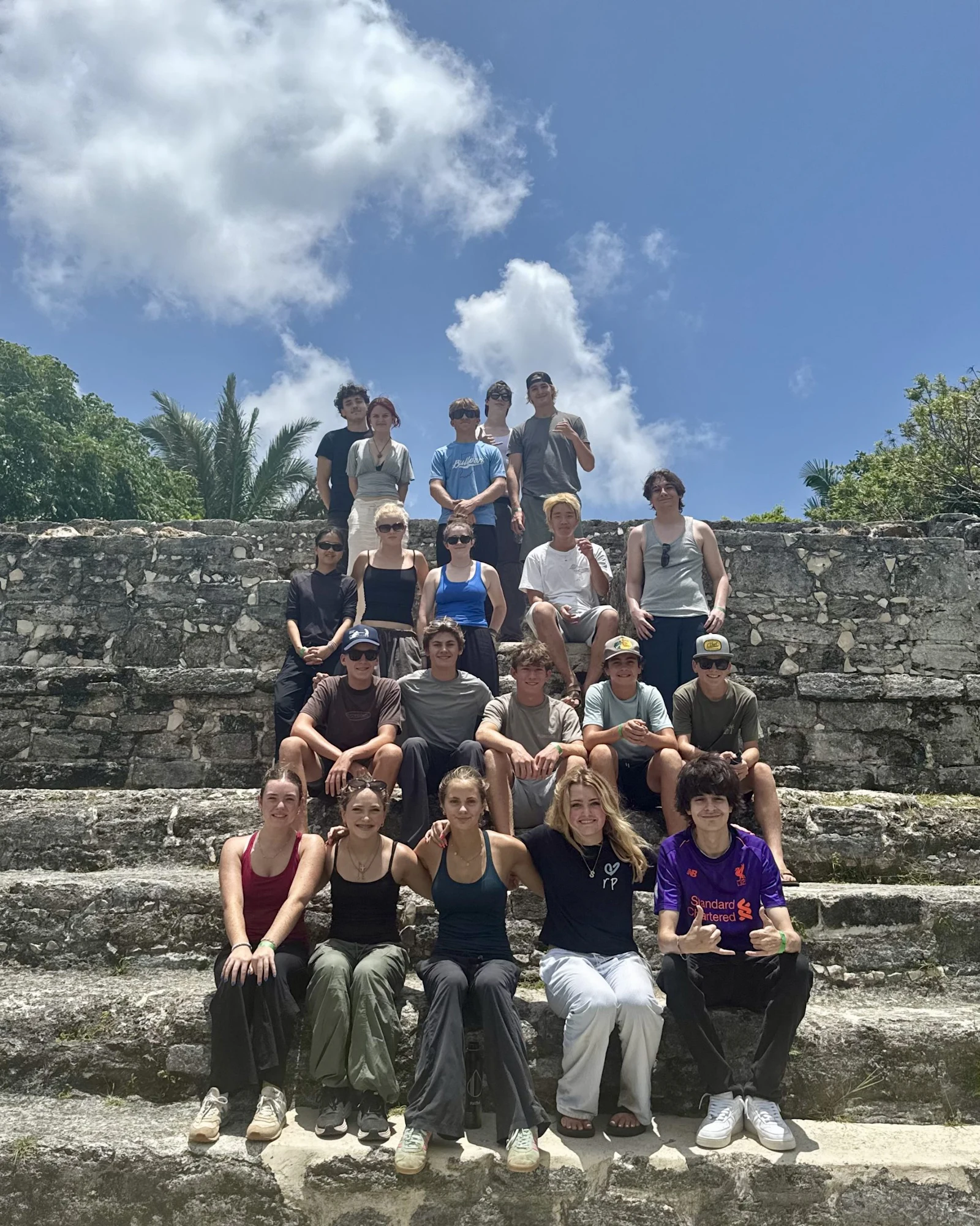 About twenty teen travelers sit and stand together on the weathered limestone steps of a Maya temple platform in Belize under a bright sky.