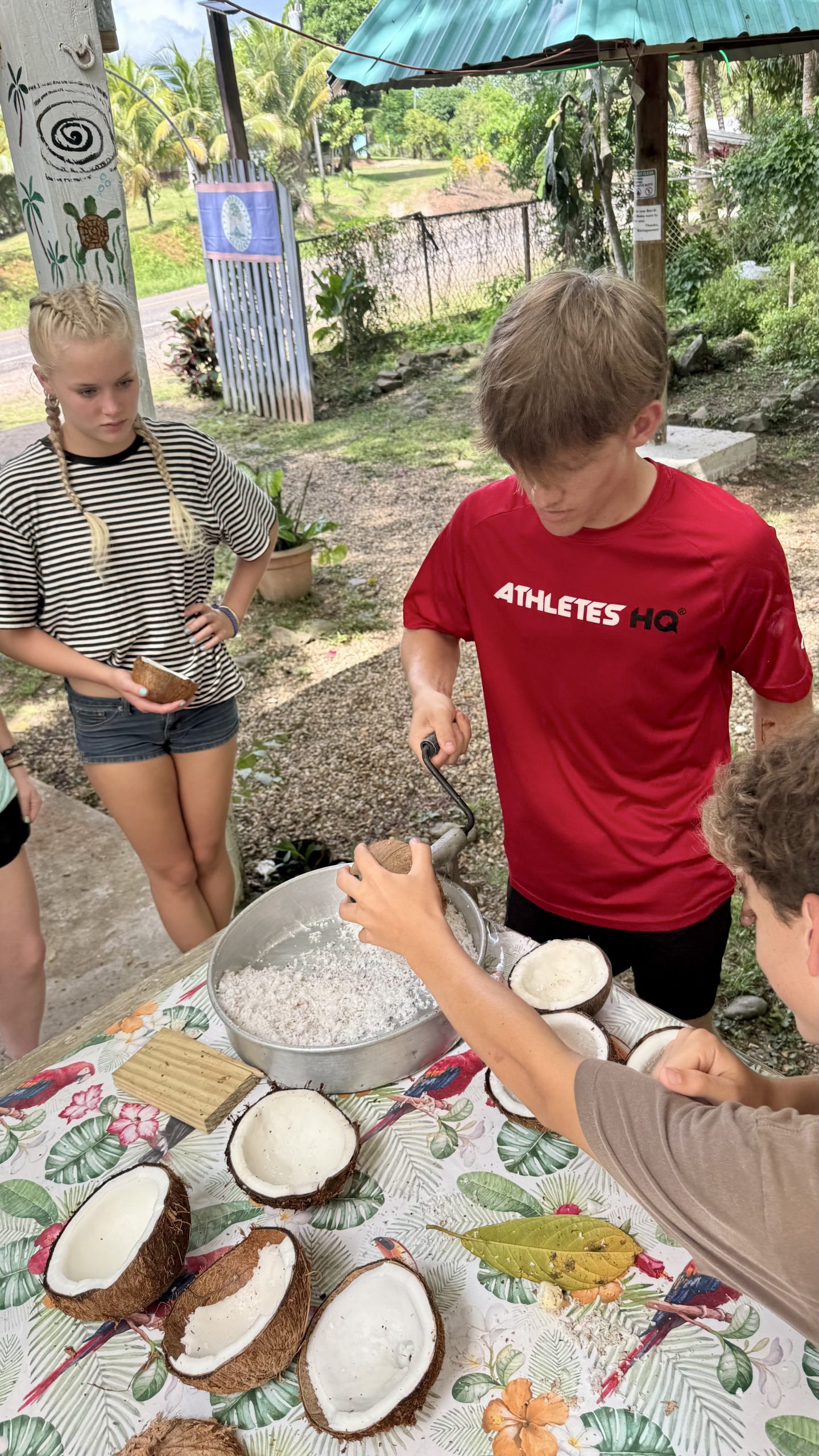 A student turns a hand-cranked grater over a metal bowl of shredded coconut during a cooking workshop in Belize.