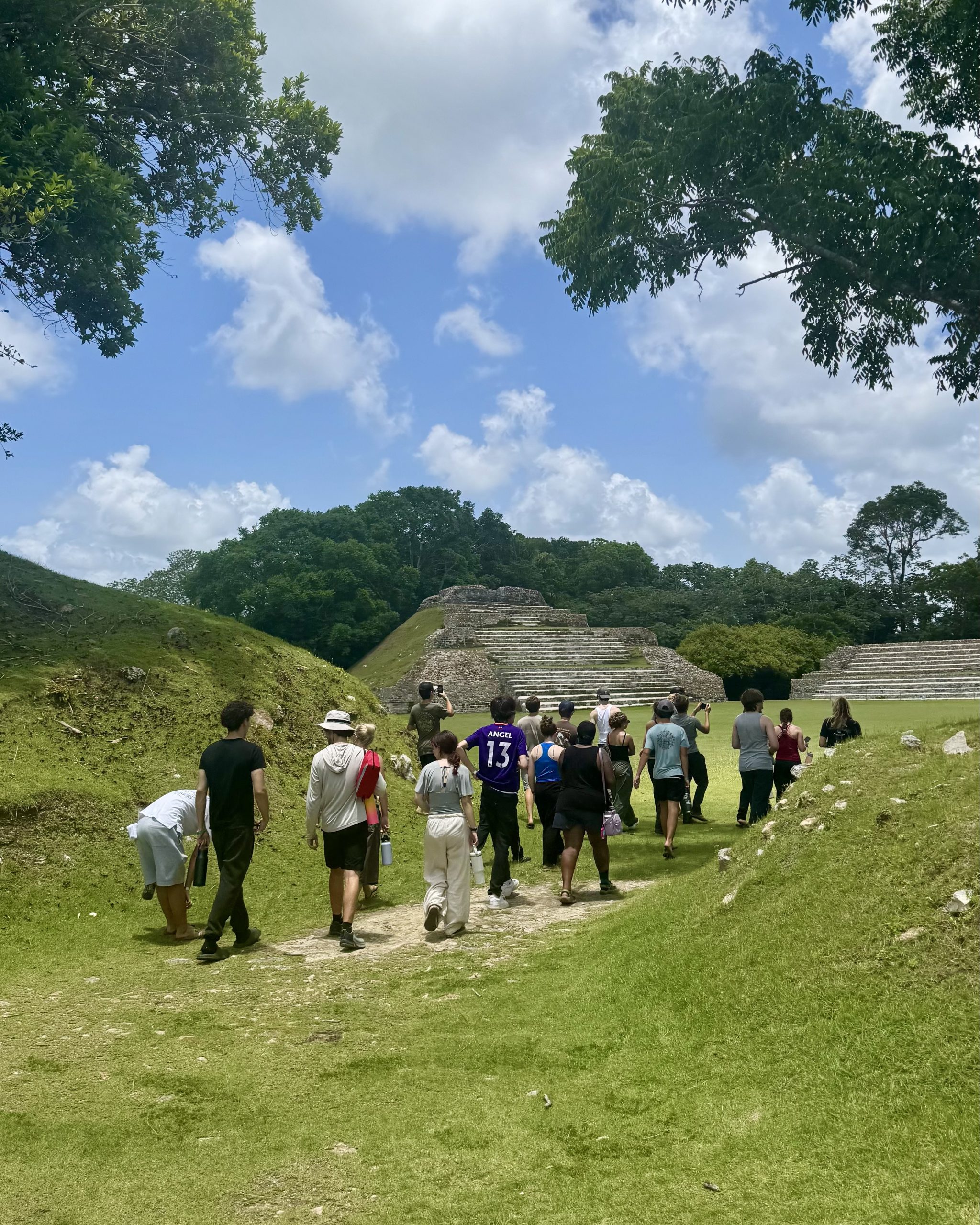 A line of teen travelers walks a grass path across a Maya site in Belize toward a stepped stone pyramid.