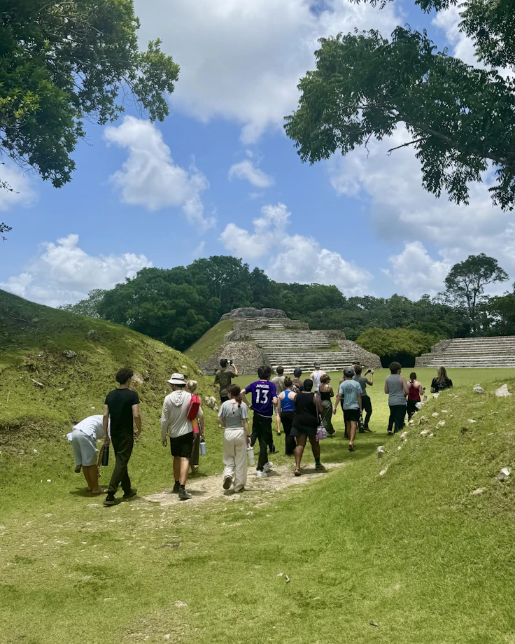 A line of teen travelers walks across a Maya site in Belize toward a stepped stone pyramid
