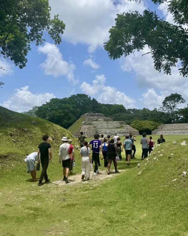 A line of teen travelers walks along a grass path across a Maya site in Belize toward a stepped stone pyramid framed by jungle
