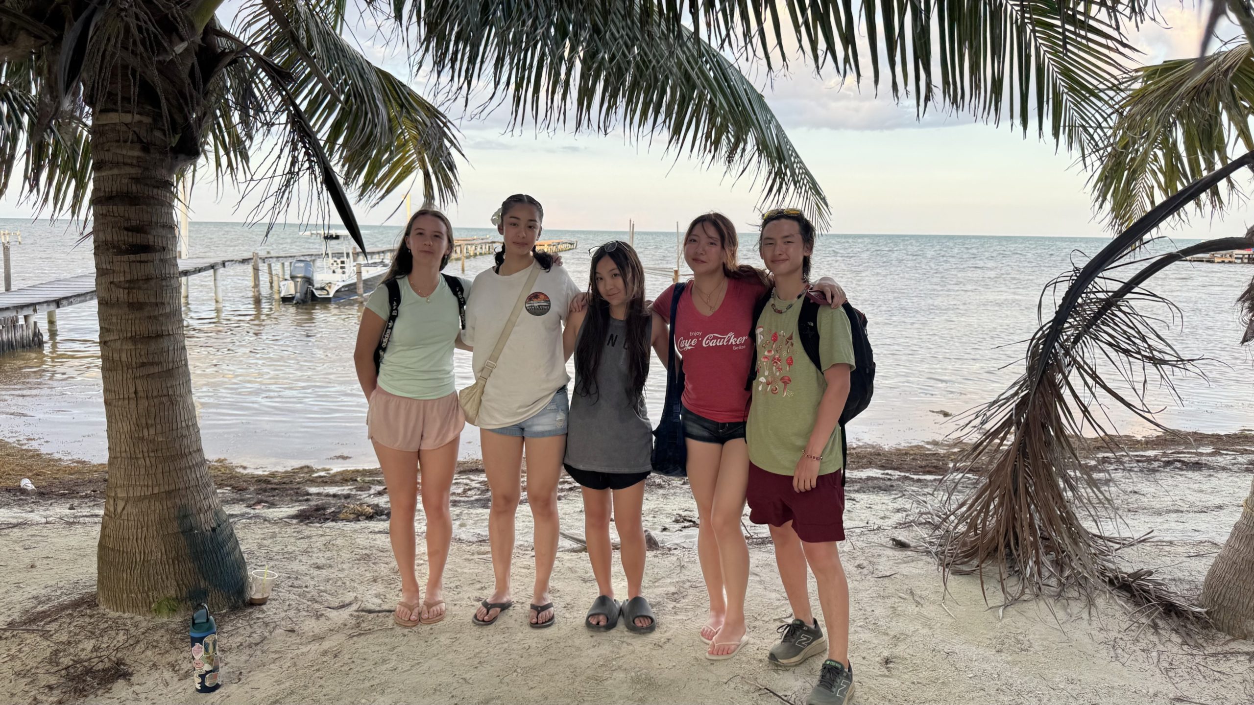 Teens pose beneath palm trees on a beach with a dock and calm water behind them