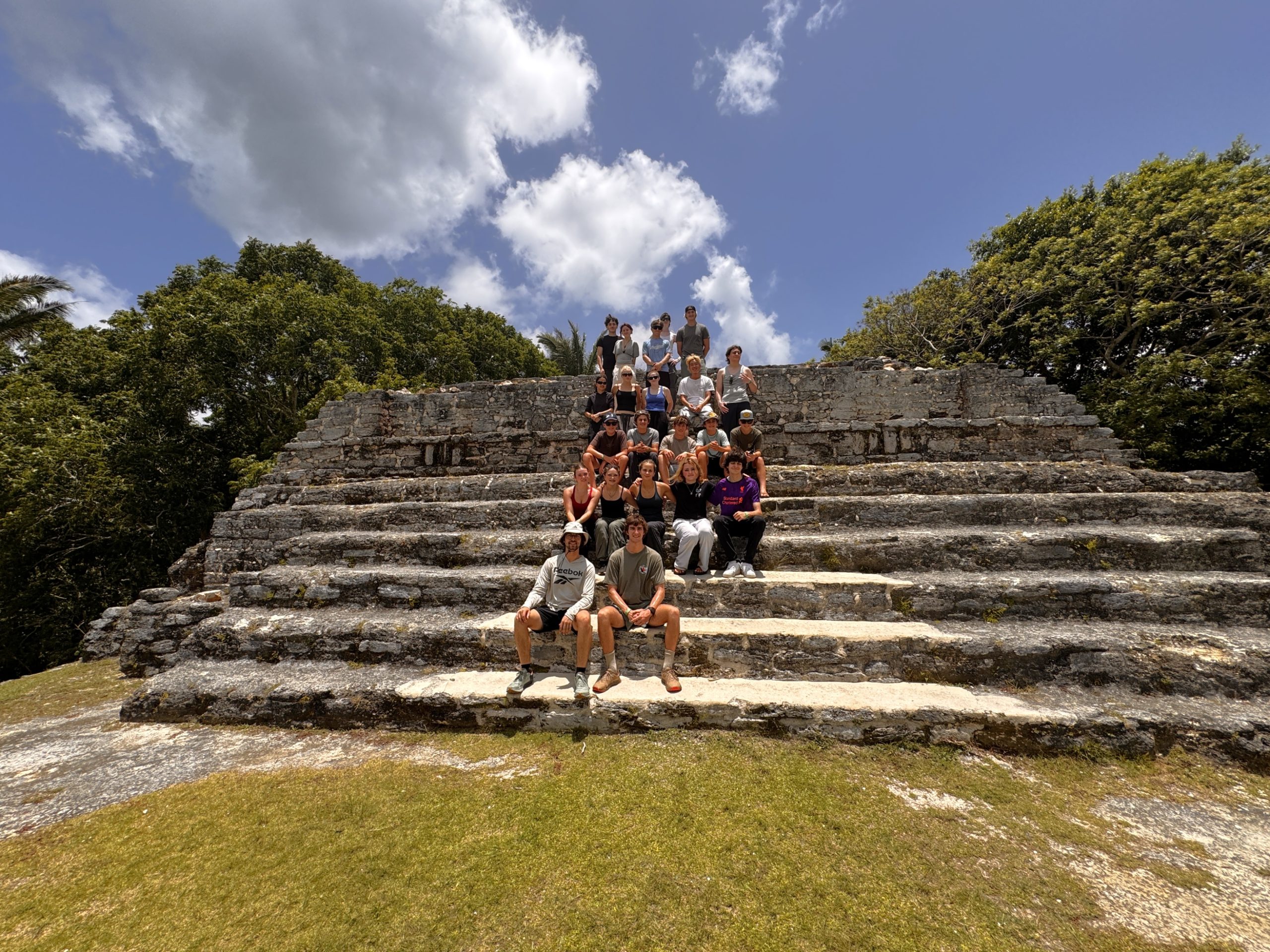 Student travelers sit together on the stone steps of a Maya pyramid under a blue sky