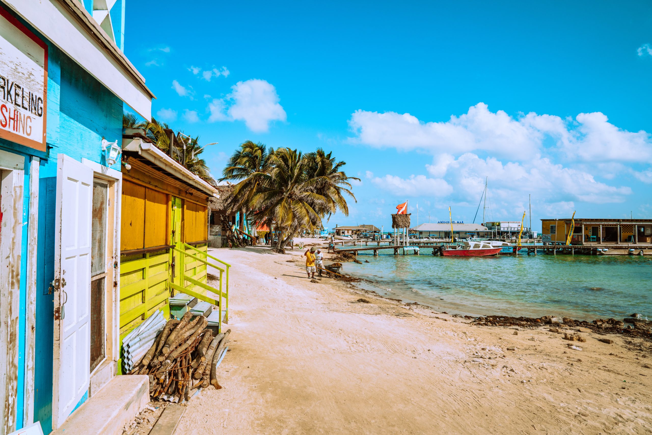 Sandy beachfront path in Belize lined with colorful buildings and palm trees leads to a dock with boats